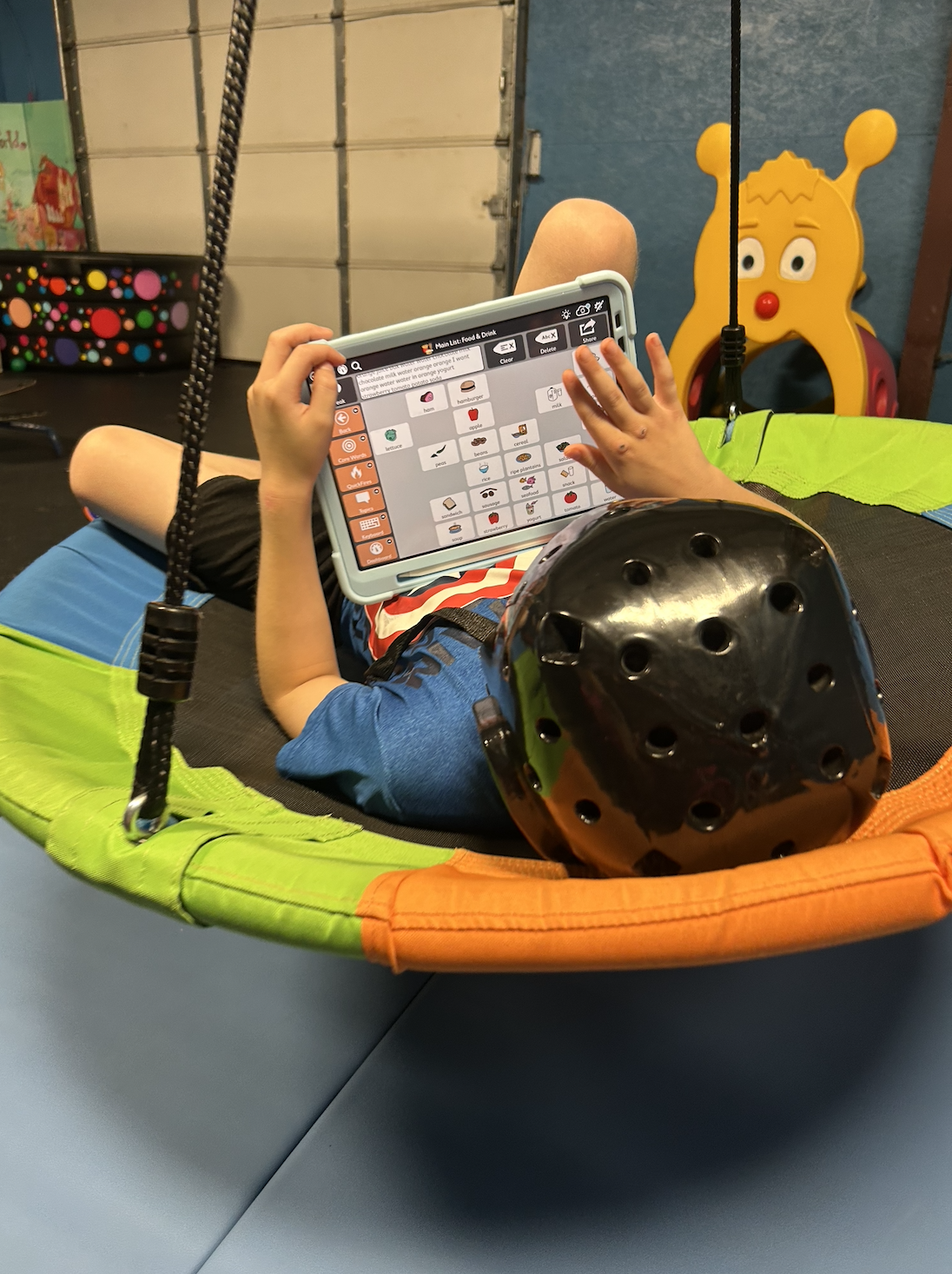 A child lying on a colorful swingset with a toy helmet, holding a tablet with an app displaying food and drink icons, in a play area with a toy giraffe in the background.