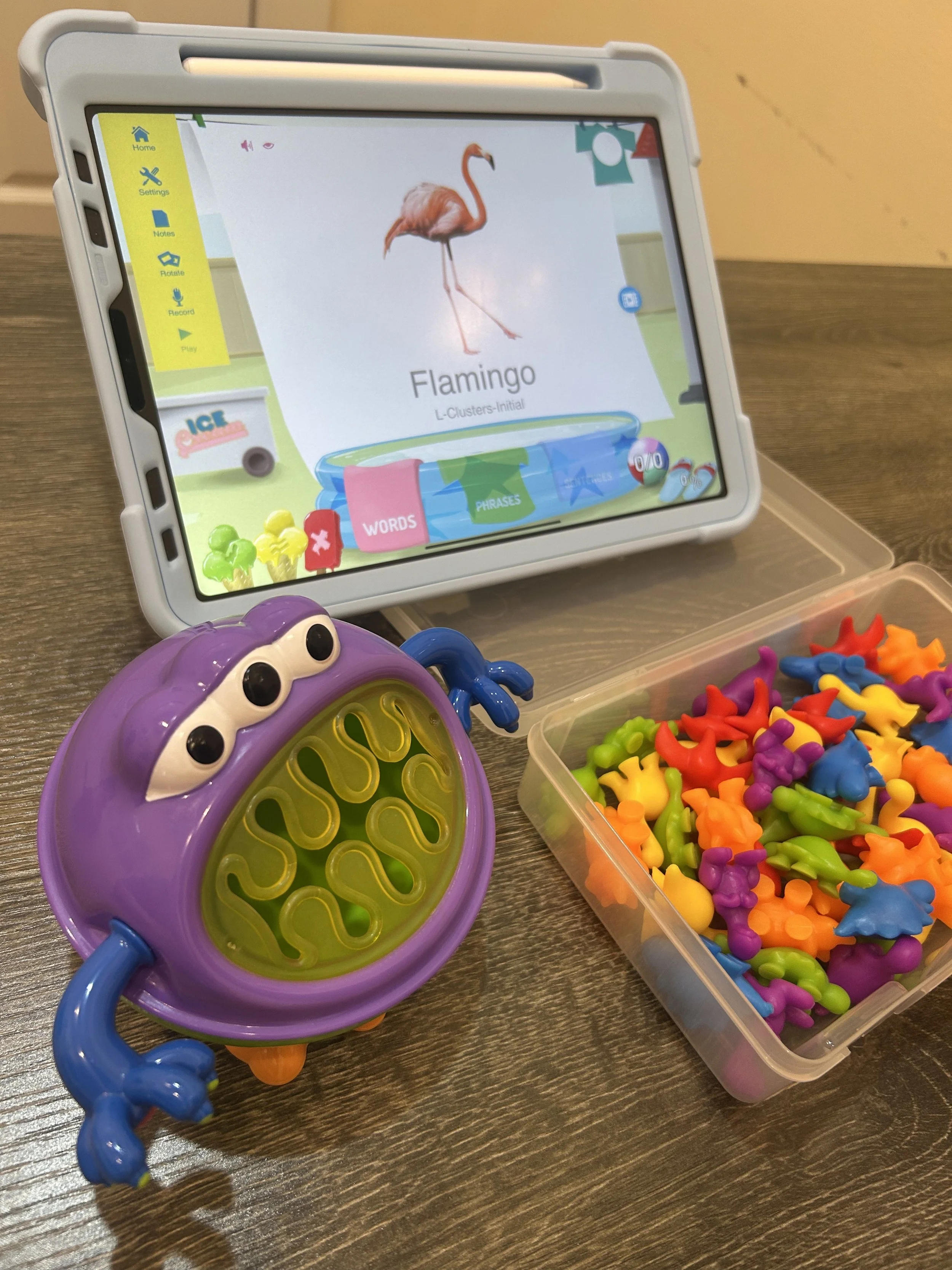 An educational activity setup showing a tablet displaying a picture of a flamingo with the label 'Flamingo' underneath, a purple bug-shaped word game with a yellow interior, and a transparent container filled with colorful plastic dinosaur-shaped tokens.