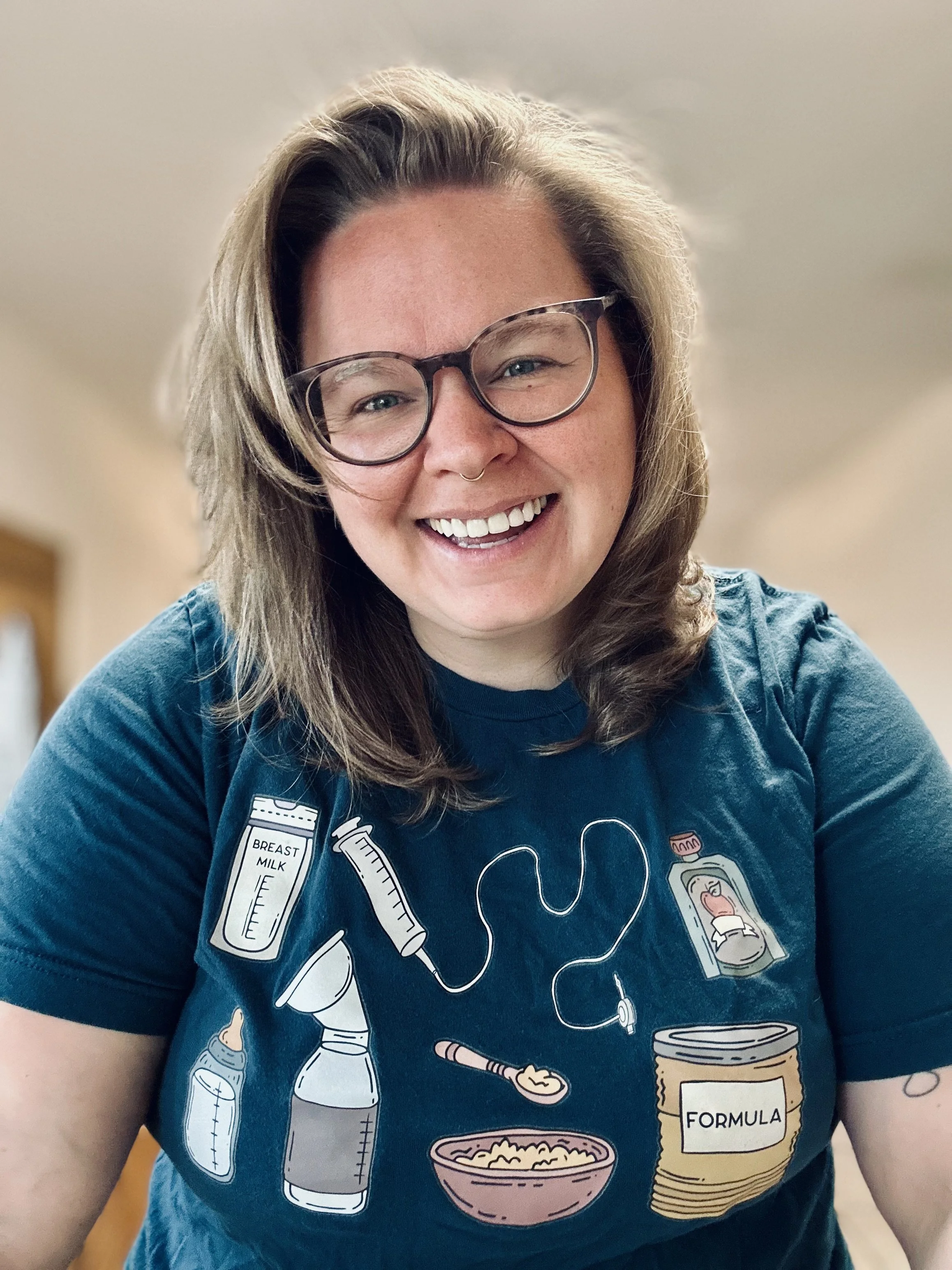 A smiling woman with glasses and a septum piercing wearing a blue t-shirt with illustrations of milk, a syringe, a stethoscope, a baby bottle, a spoon, a bowl, and formula.
