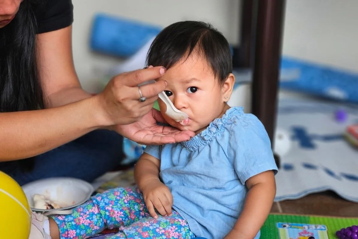 A young child with short dark hair and earrings, sitting on a mat, is being fed with a spoon by an adult. The child is wearing a light blue dress and colorful floral pants. The background includes a bed with blue bedding and some toys.
