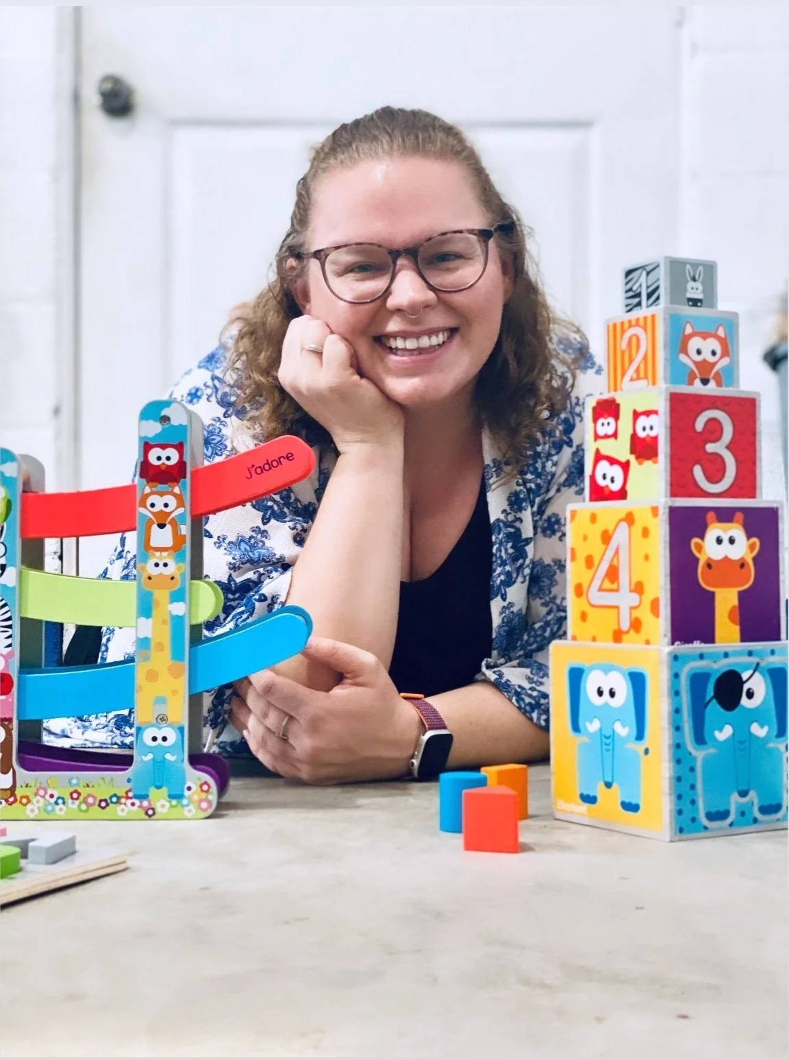 A woman with glasses and a flower-patterned shirt sitting at a table with children's toys, smiling at the camera. The toys include a colorful stack of blocks with animal faces and a wooden ring stacking game, indicating a playful and cheerful environment.