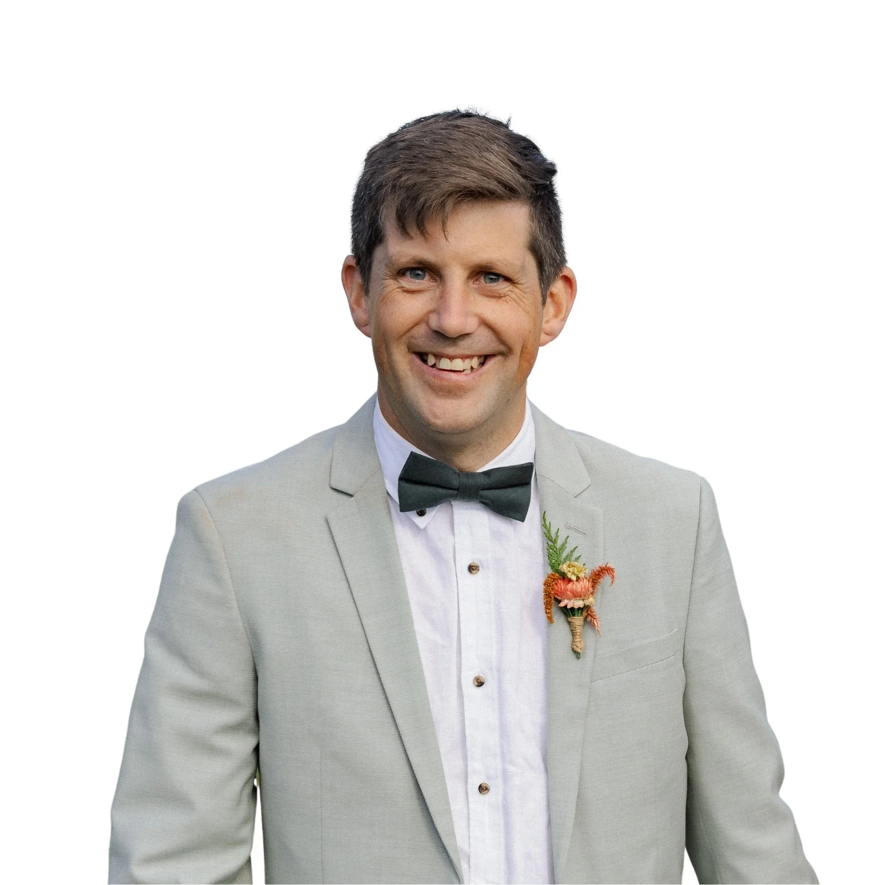 A man with brown hair wearing a light gray suit jacket, white shirt, and black bowtie, smiling, with a boutonniere pinned to his jacket lapel.