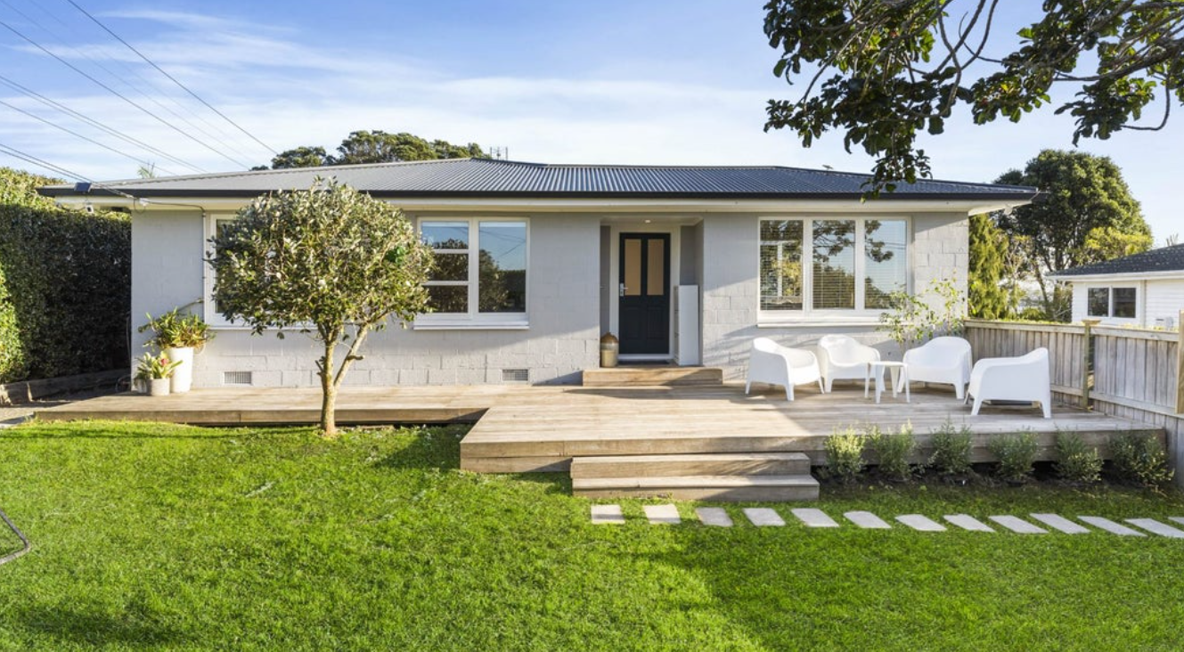 A cozy backyard with a wooden deck, white outdoor furniture, a small tree, lush green lawn, and a house with white walls and big windows.