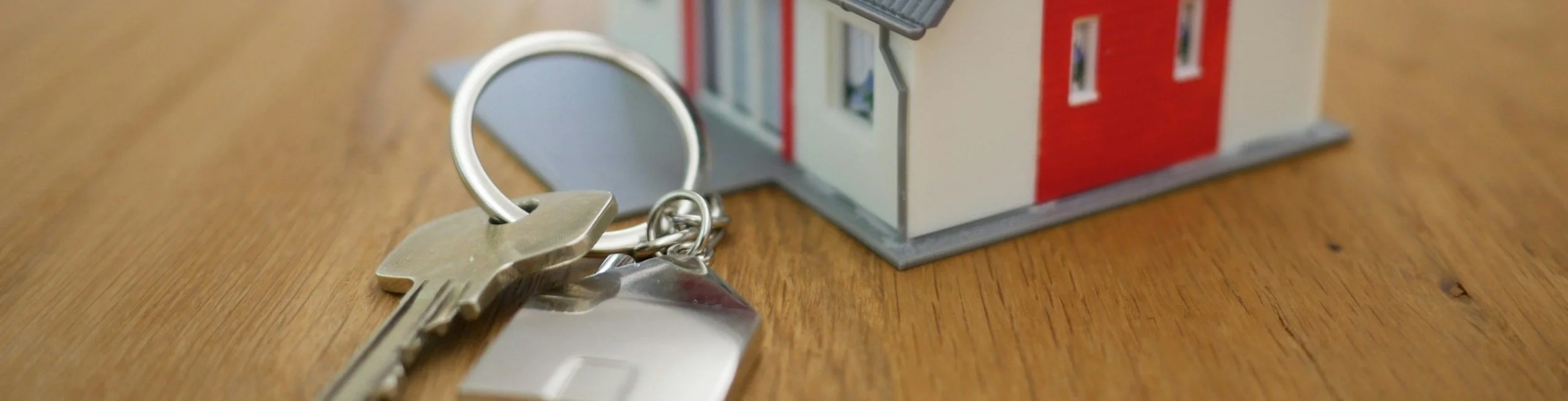 Keys on a keyring placed on a wooden surface next to a small model house.