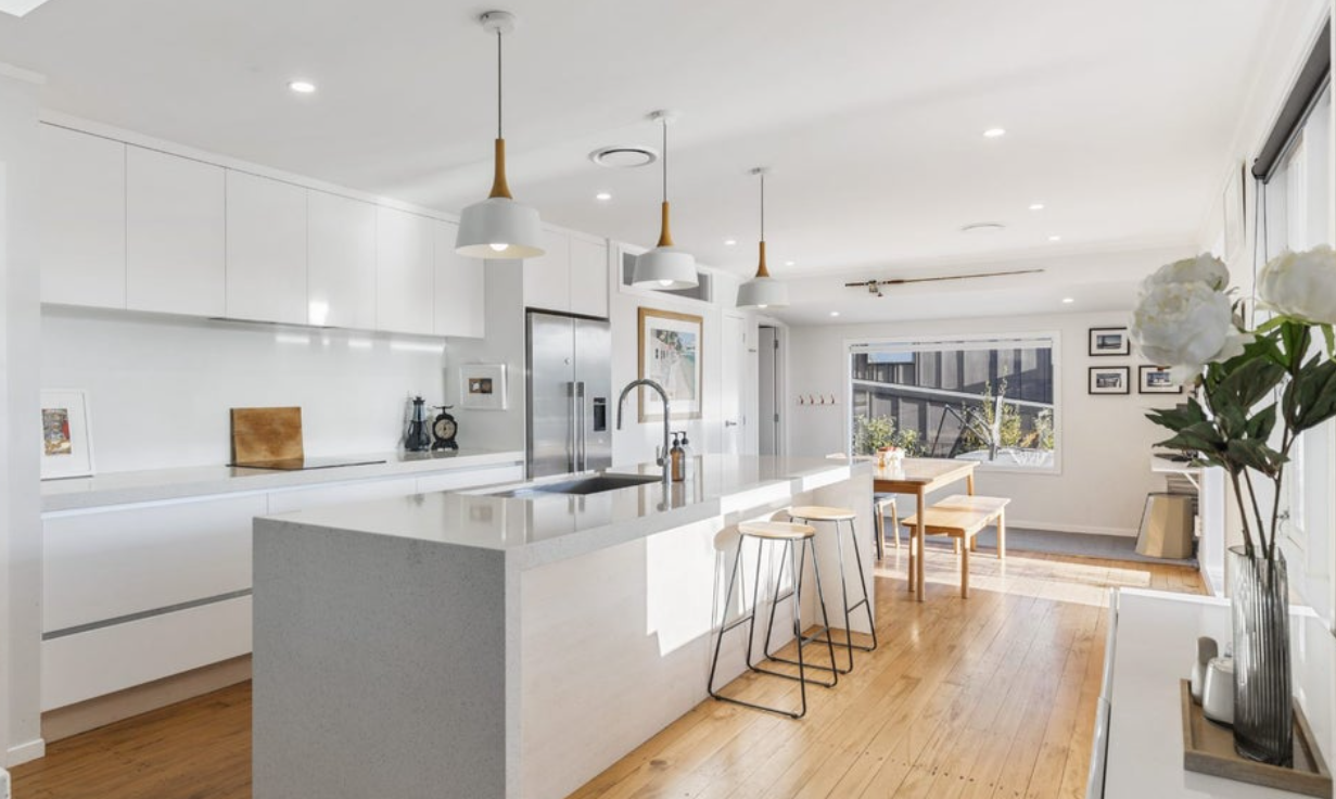 Bright modern kitchen with white cabinets, island with seating, and wooden flooring, overlooking a dining area and large window