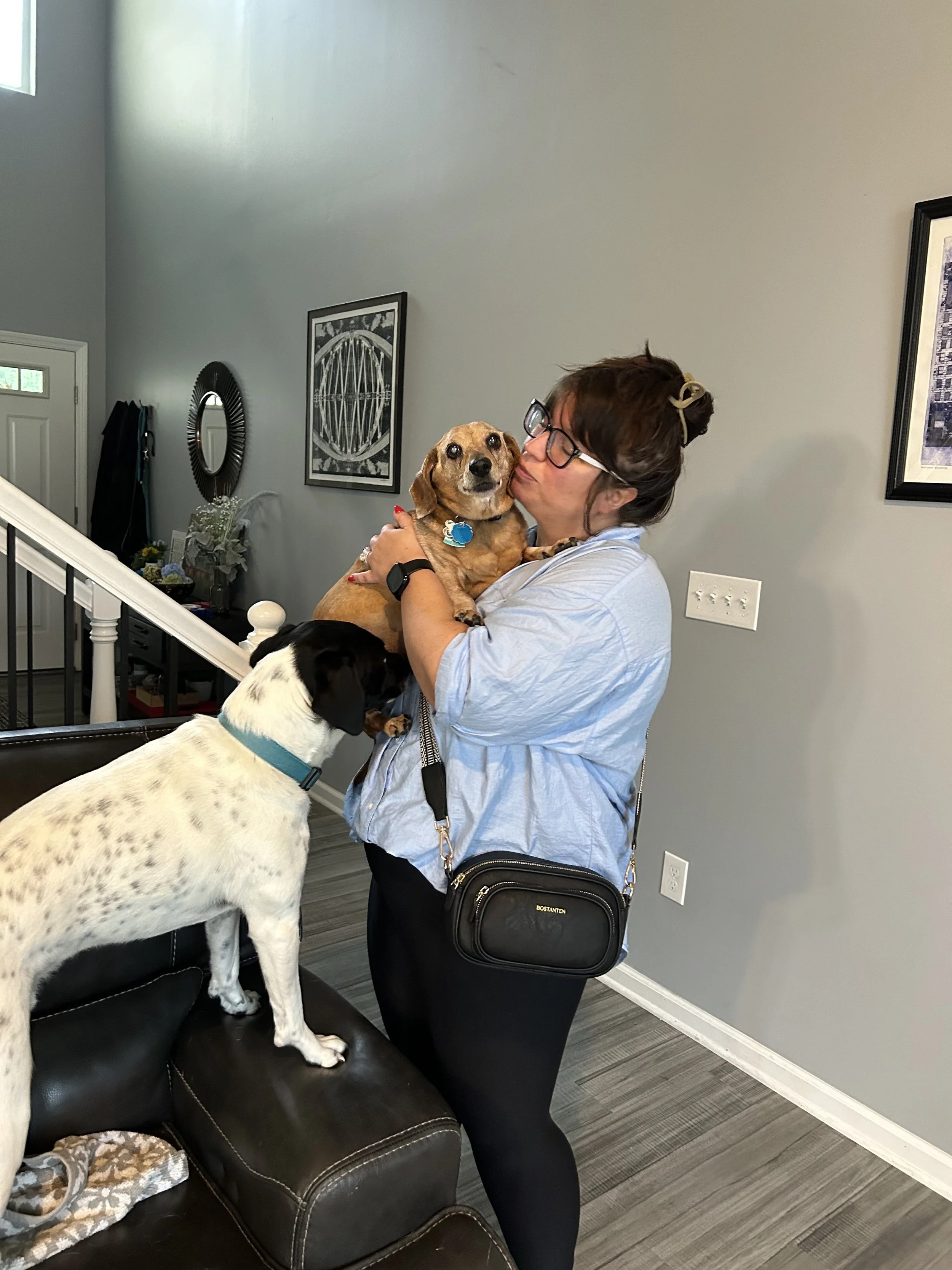 A woman is hugging and kissing a small dog while a large dog stands on a sofa, inside a living room.