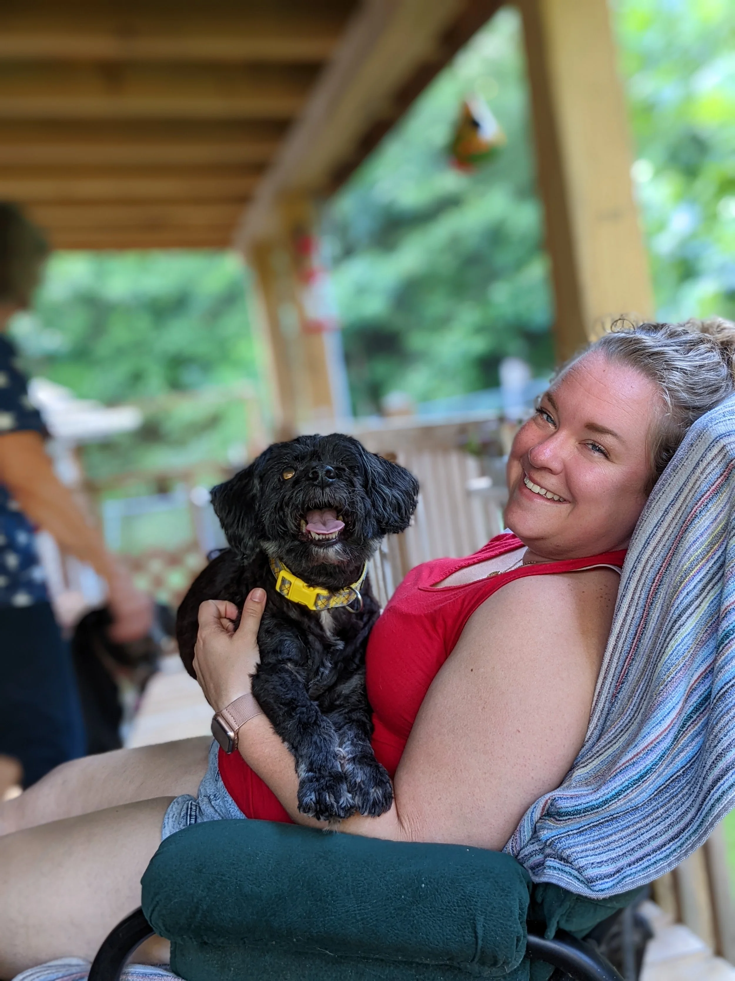 A woman smiling and sitting on a porch, holding a small black dog with a yellow collar. The porch has wooden beams and a blurred background of green trees.