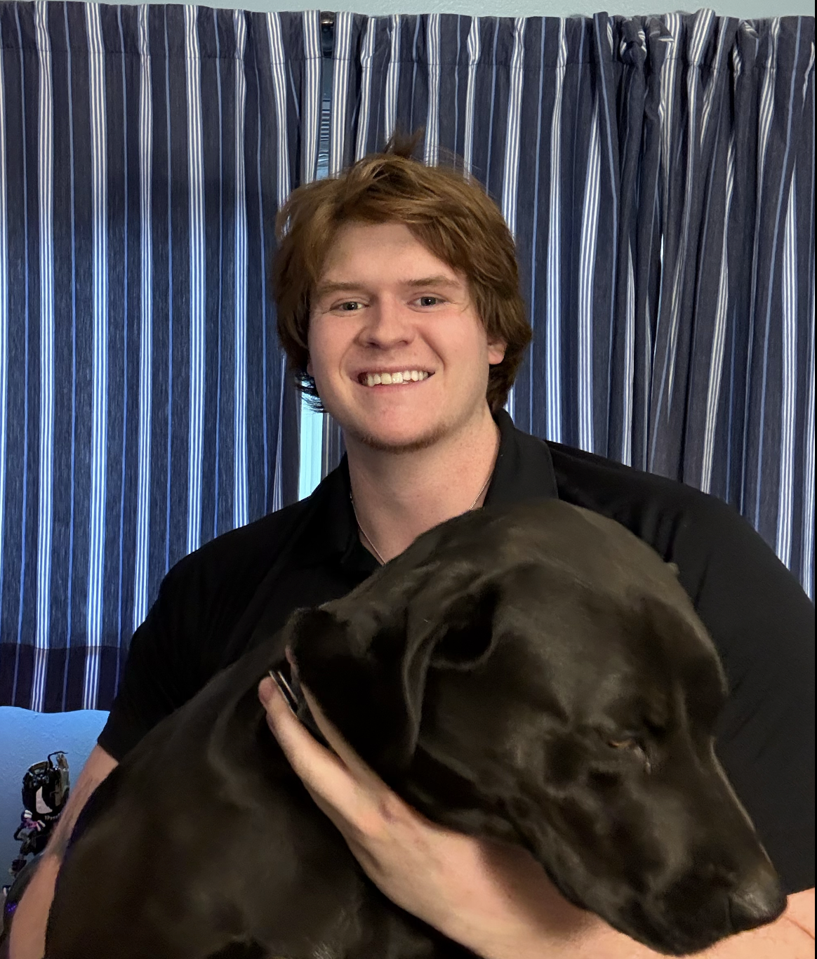 A young man with red hair smiling and holding a black dog with floppy ears in front of blue striped curtains.