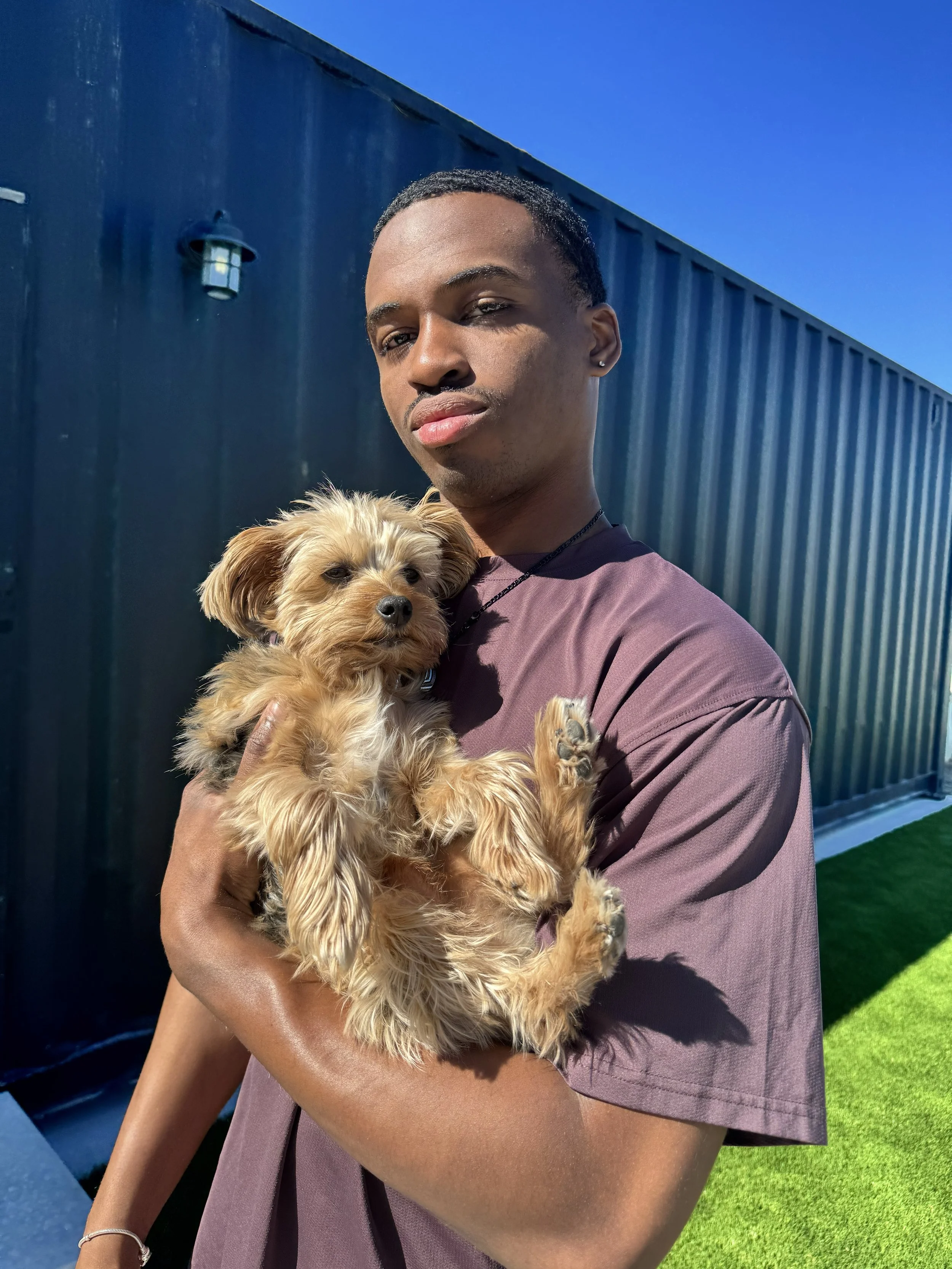 A young man holding a small, fluffy, light brown dog outdoors in front of a dark-colored wall and a blue sky.
