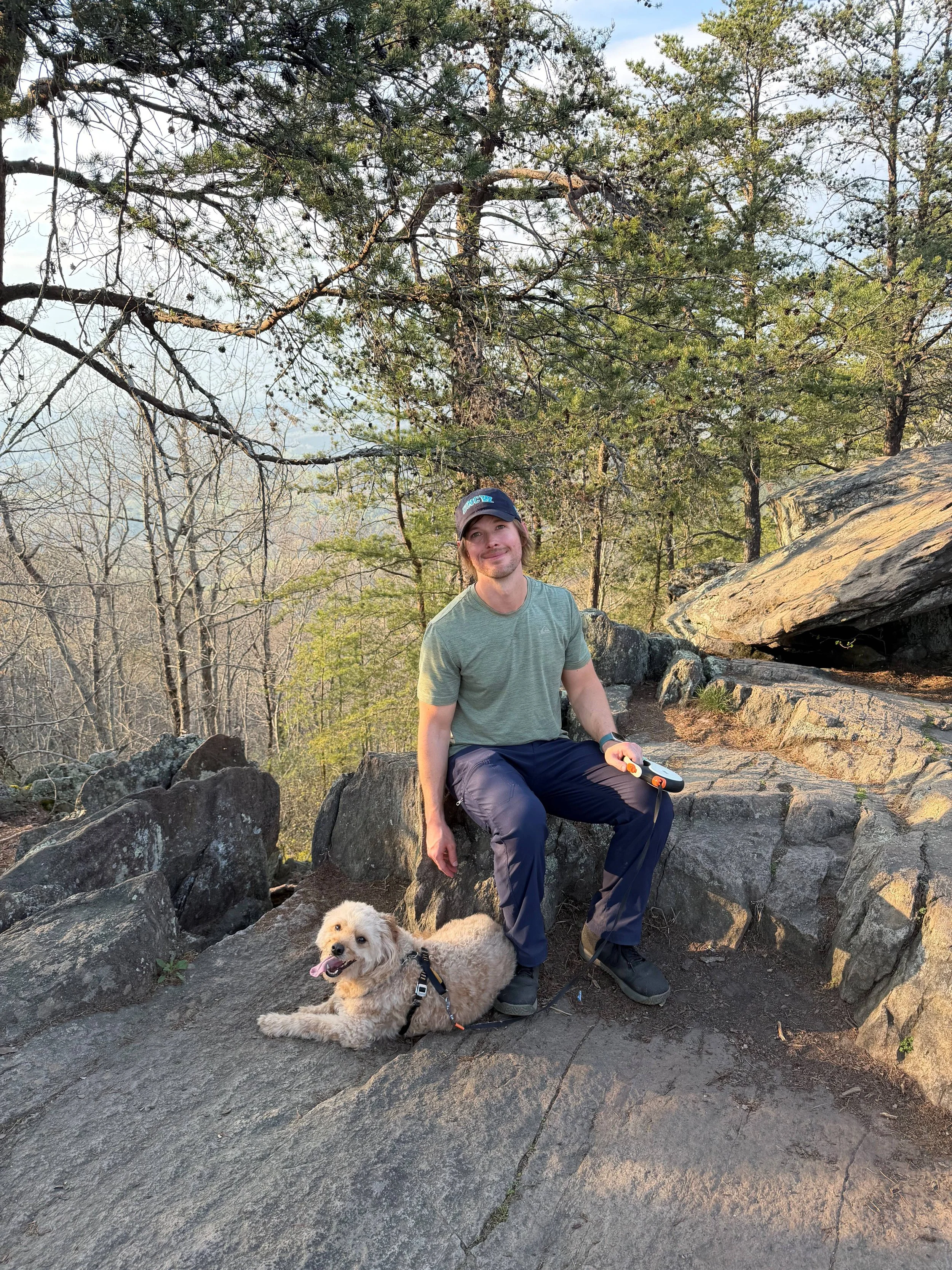 A man sitting on a rock with a dog lying next to him in a forested outdoor setting, with trees and rocks in the background.