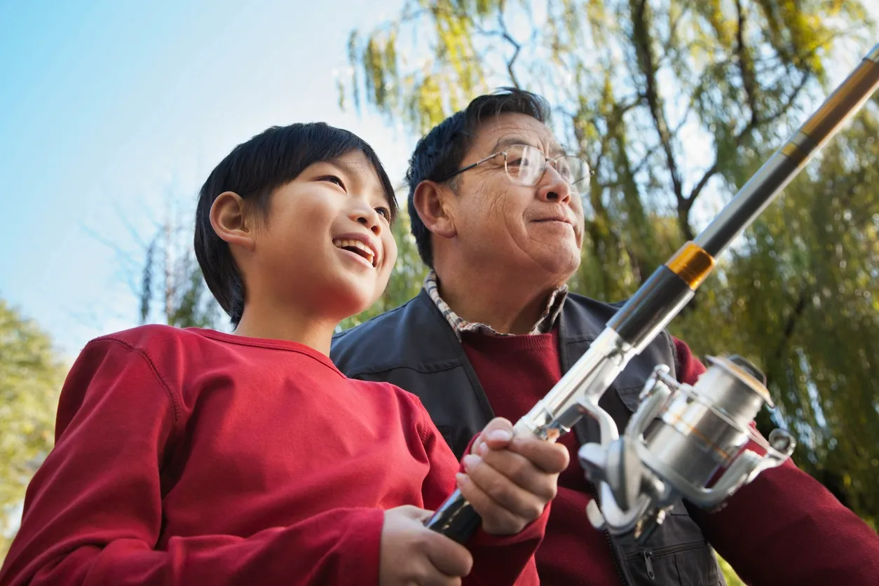 A young boy and an older man, possibly his grandfather, outdoors in a wooded area, enjoying fishing together. The boy is holding a fishing rod and smiling, while the man looks on with a proud expression.