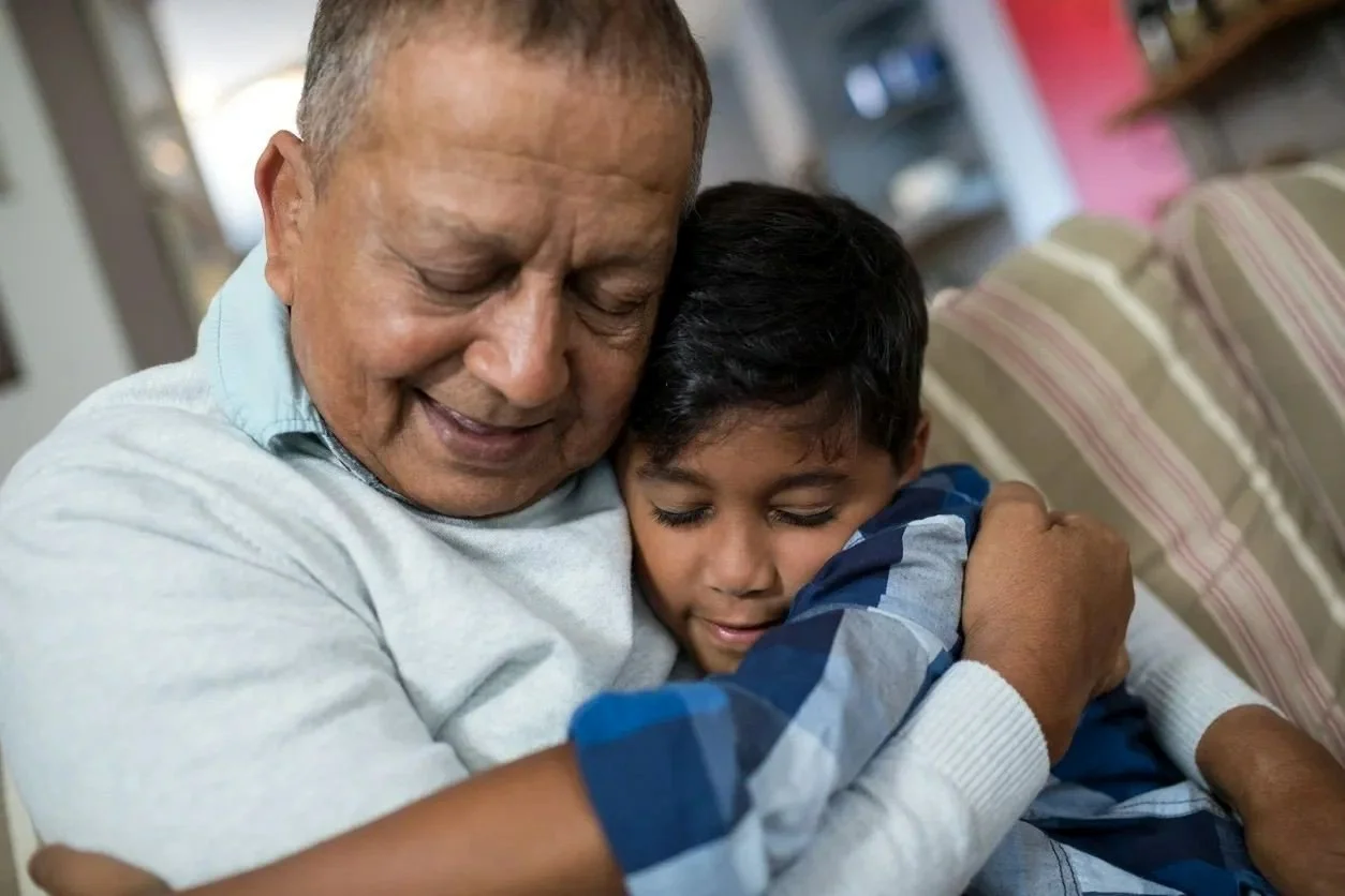 An elderly man and a young boy are hugging each other tightly and both have their eyes closed, expressing warmth and love in a cozy indoor setting.
