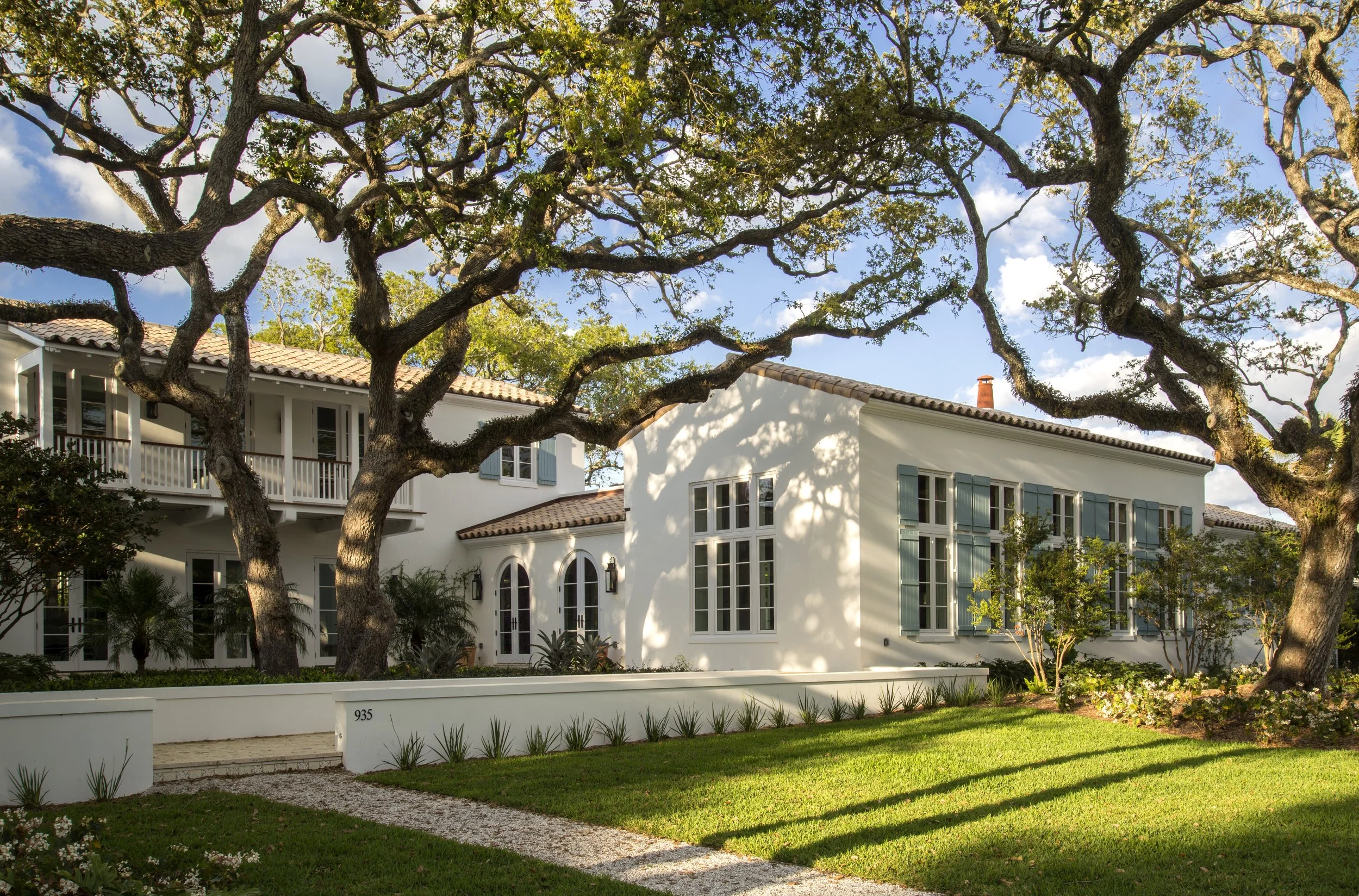 White house with a gray tiled roof, two palm trees in front, and a brick driveway under a clear blue sky.