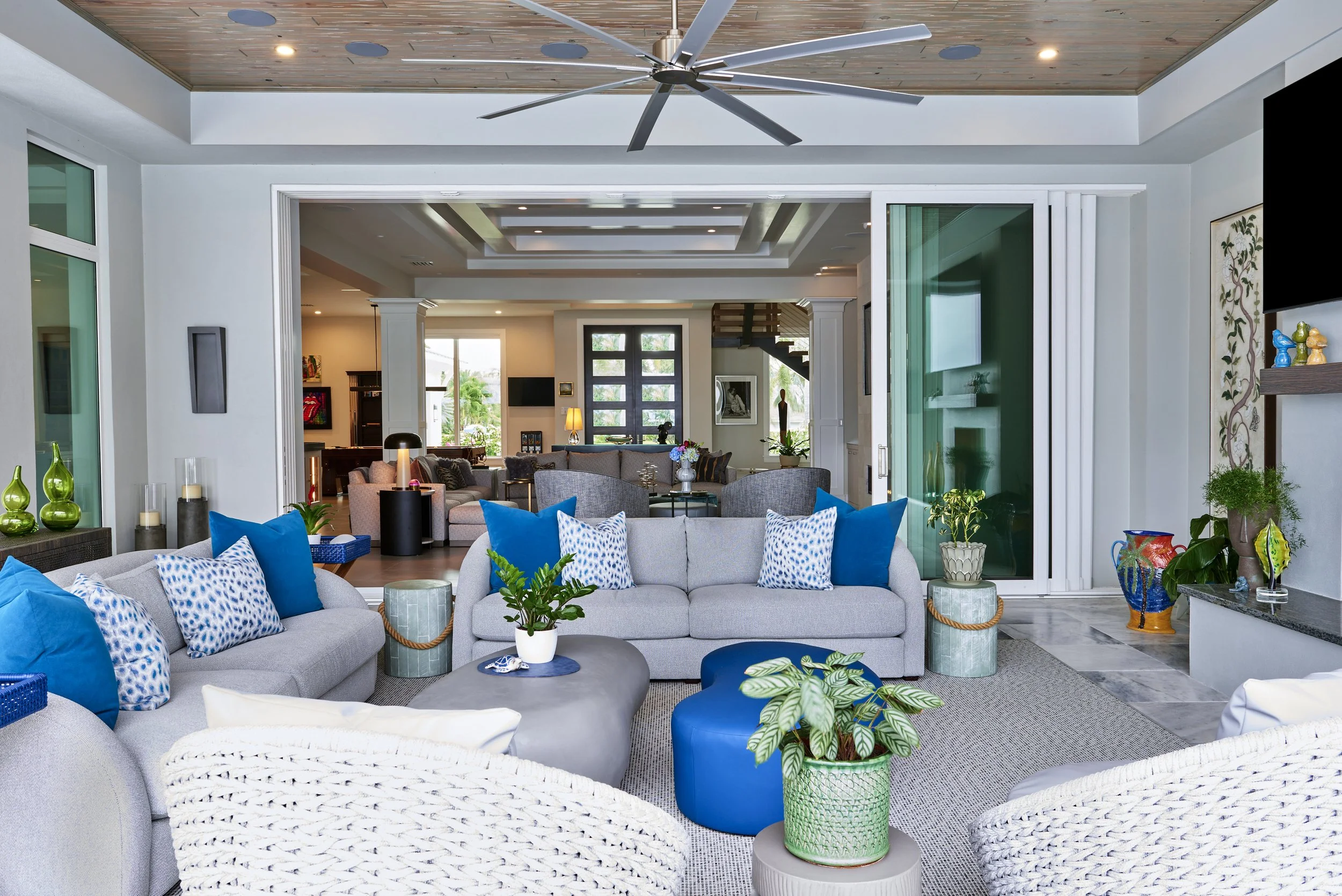 Spacious living room with white walls, hardwood floors, and large windows showing greenery outside. Light-colored sofa and chairs with decorative pillows, a dark coffee table with books and plants, and a side table with a lamp. Open kitchen and dining area in the background, with a TV and various decor pieces.