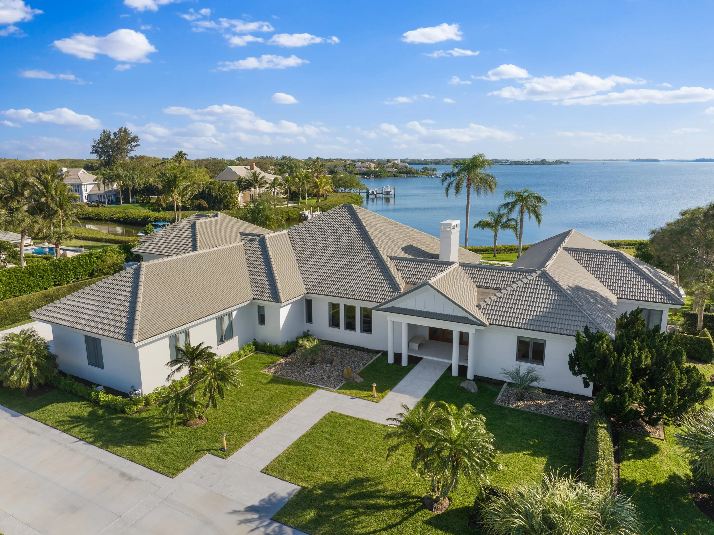 White house with a gray tiled roof, two palm trees in front, and a brick driveway under a clear blue sky.