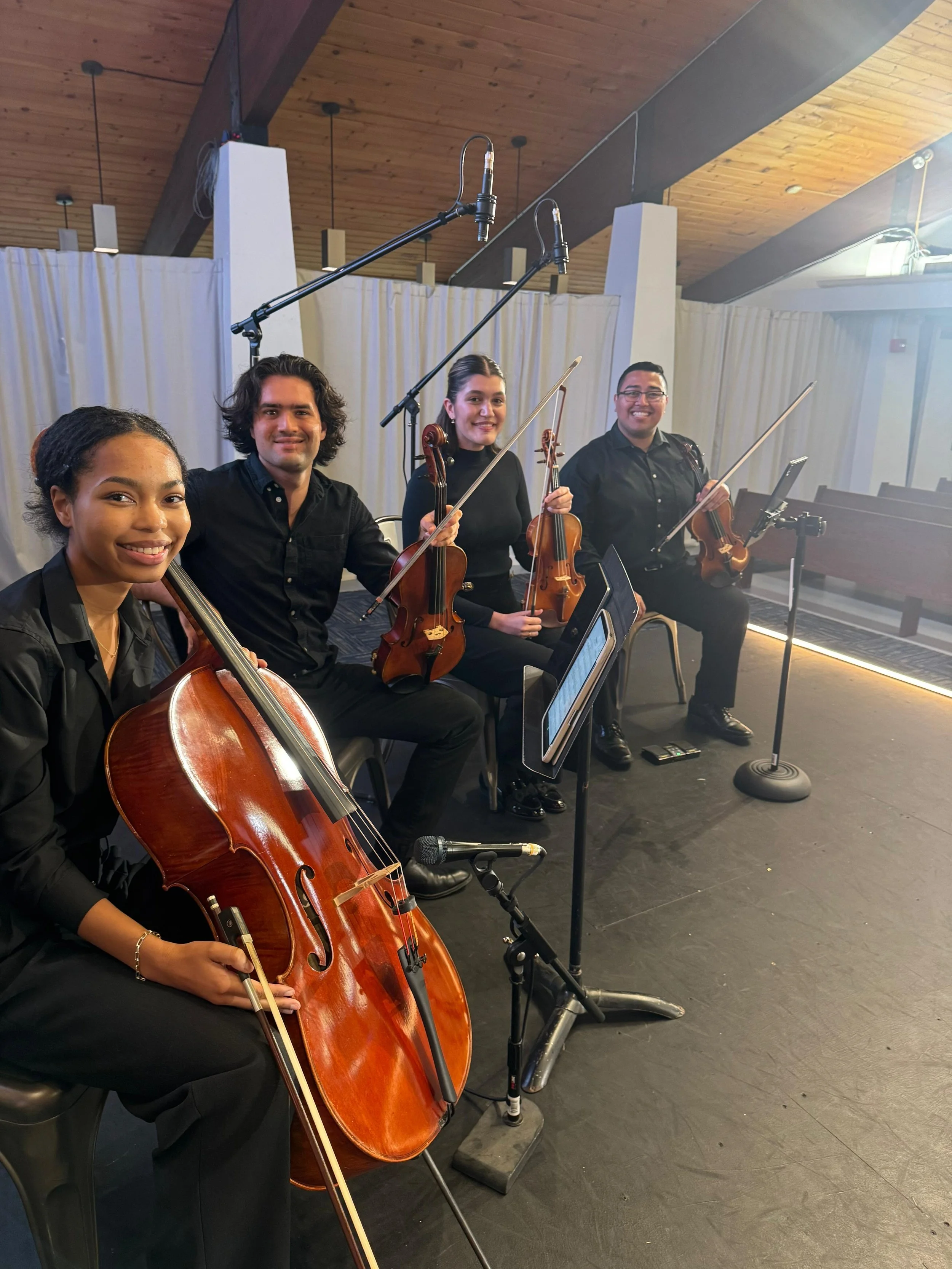 Four musicians with string instruments sitting on a stage, smiling, with music stands and microphones nearby.
