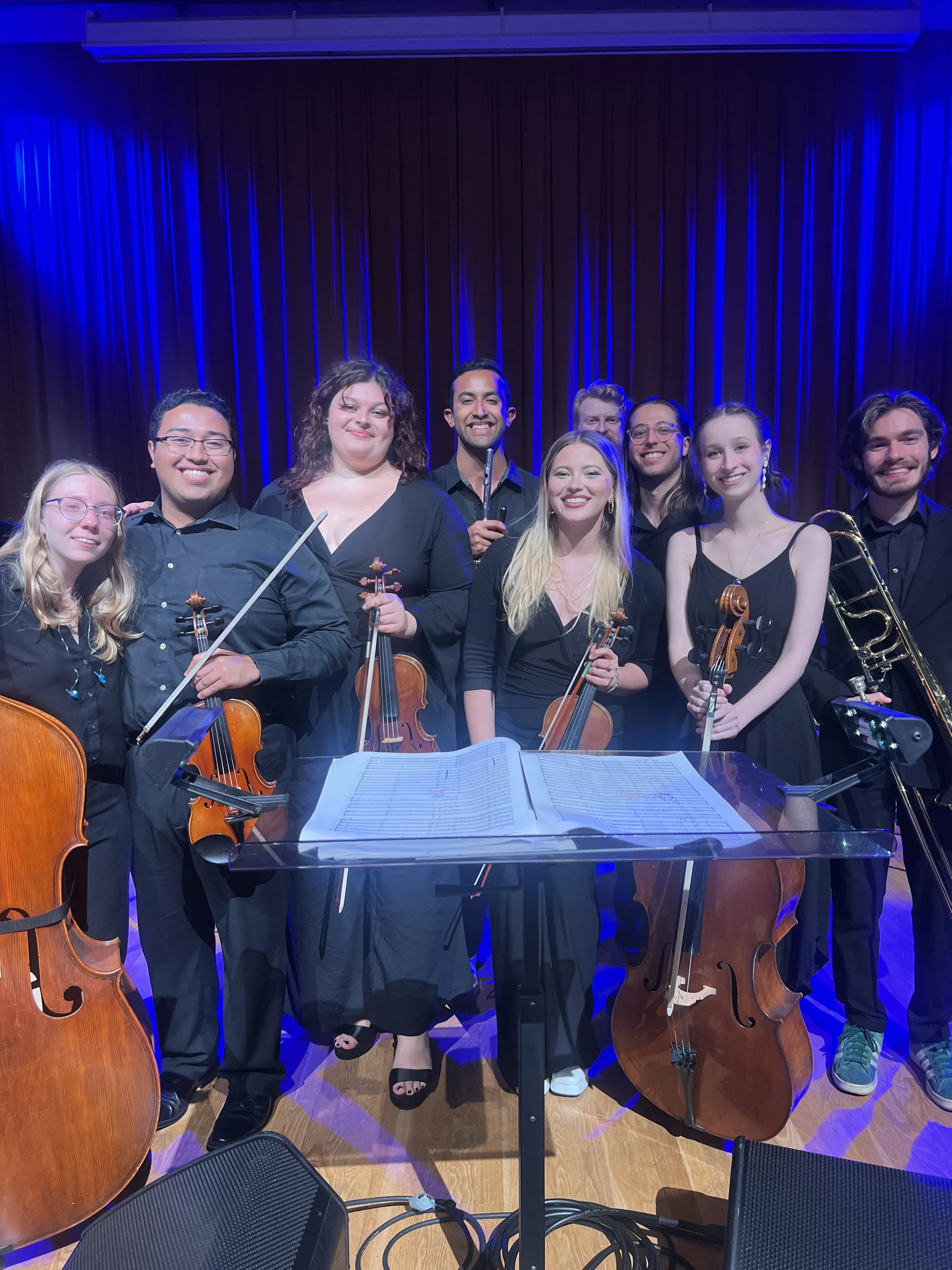 Orchestra of nine musicians standing on stage with string instruments, sheet music stand in front, dark background with blue stage lighting, posed and smiling after a performance.