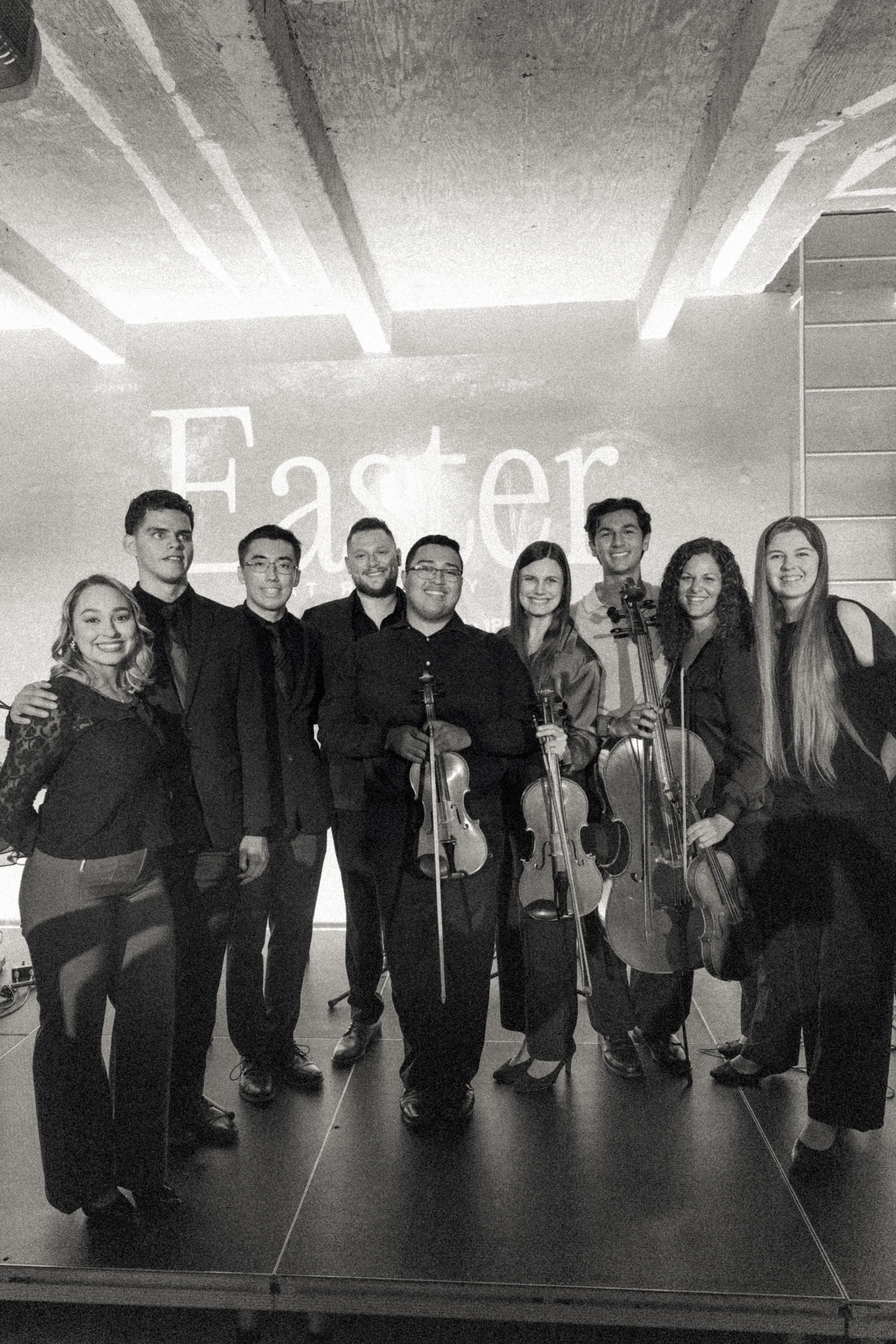 A group of nine people, including five men and four women, standing together on a stage with four of them holding violins, in front of a wall with the word 'Easter' projected on it.