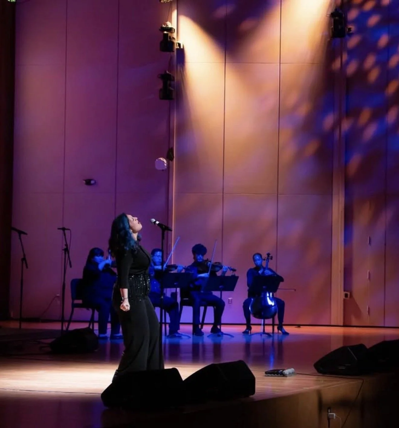 A woman singing on stage with a microphone, accompanied by a string quartet in the background, with stage lights casting colorful shadows.