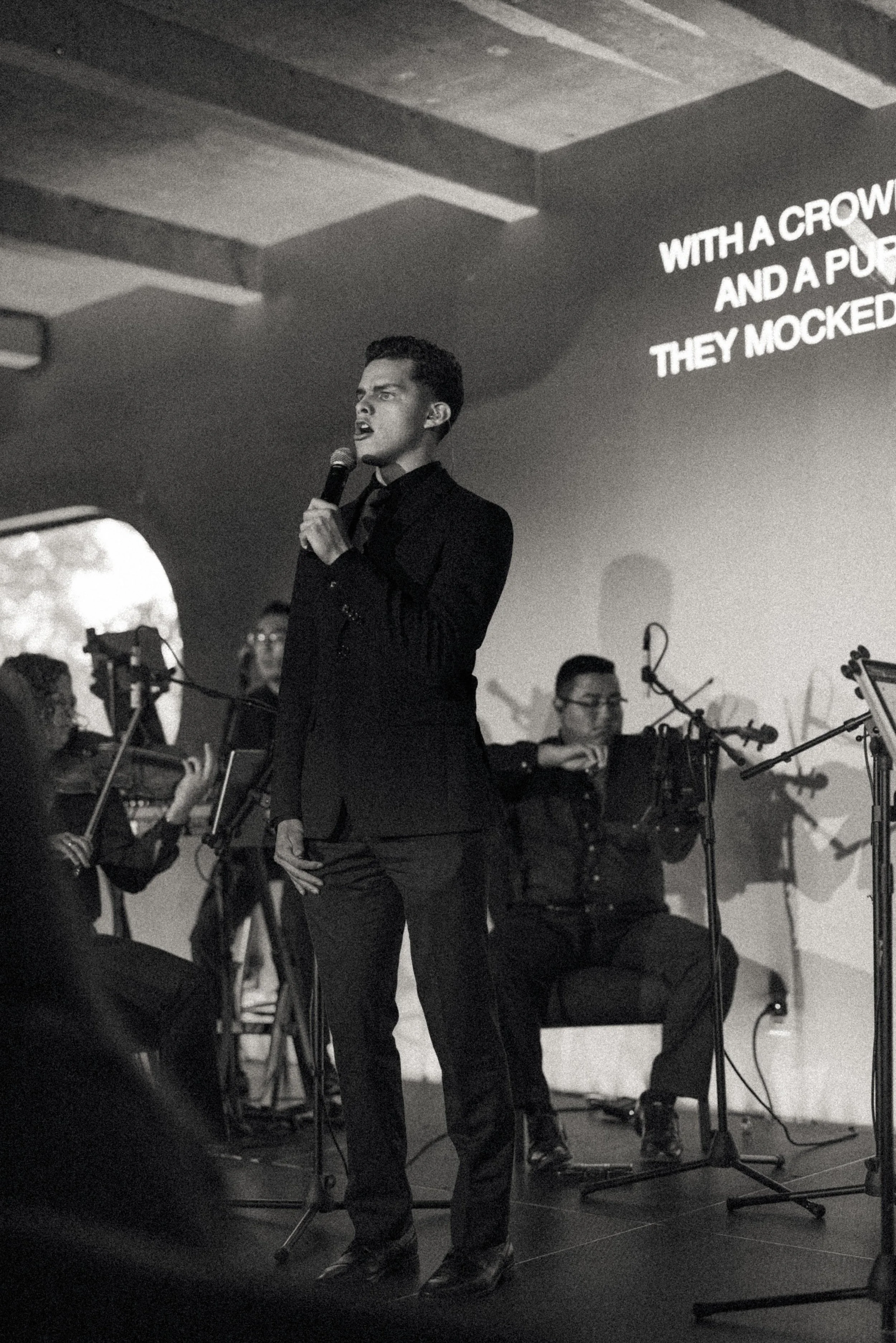 A man in a suit singing into a microphone on stage with musicians and a screen projecting lyrics in the background.