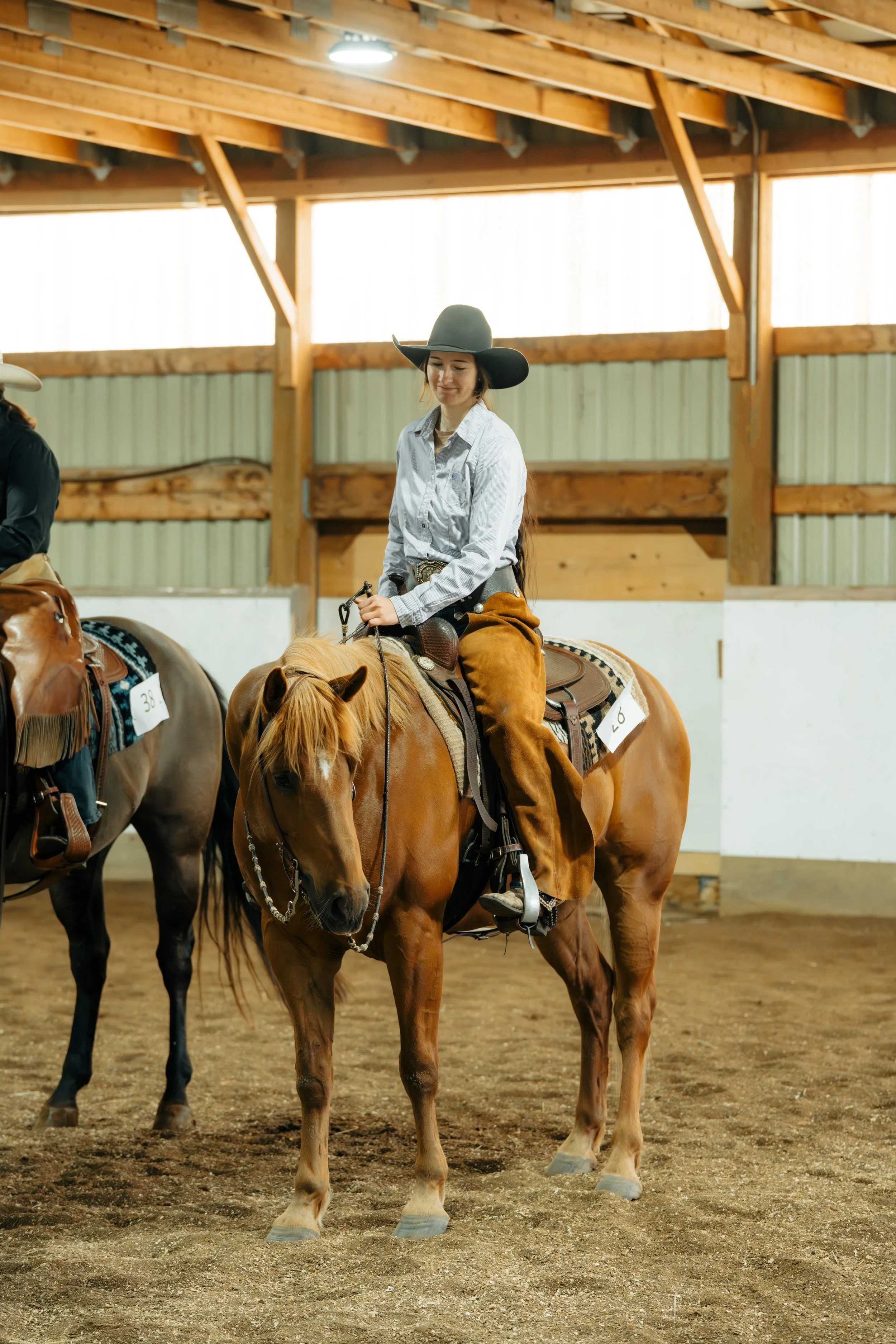 A young woman wearing a grey cowboy hat, a white button-up shirt, and brown chaps riding a light brown horse inside an indoor riding arena with wooden beams and a dirt floor.