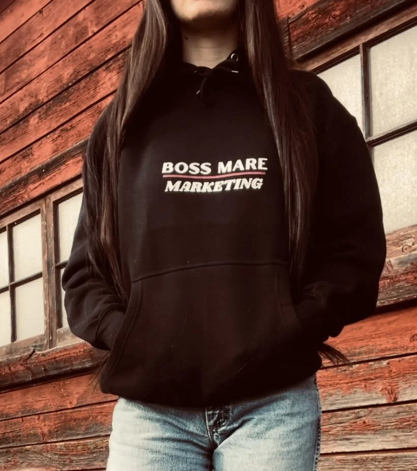 Person wearing a black hoodie with 'BOSS MARE MARKETING' printed on it, standing in front of a red wooden barn with windows.