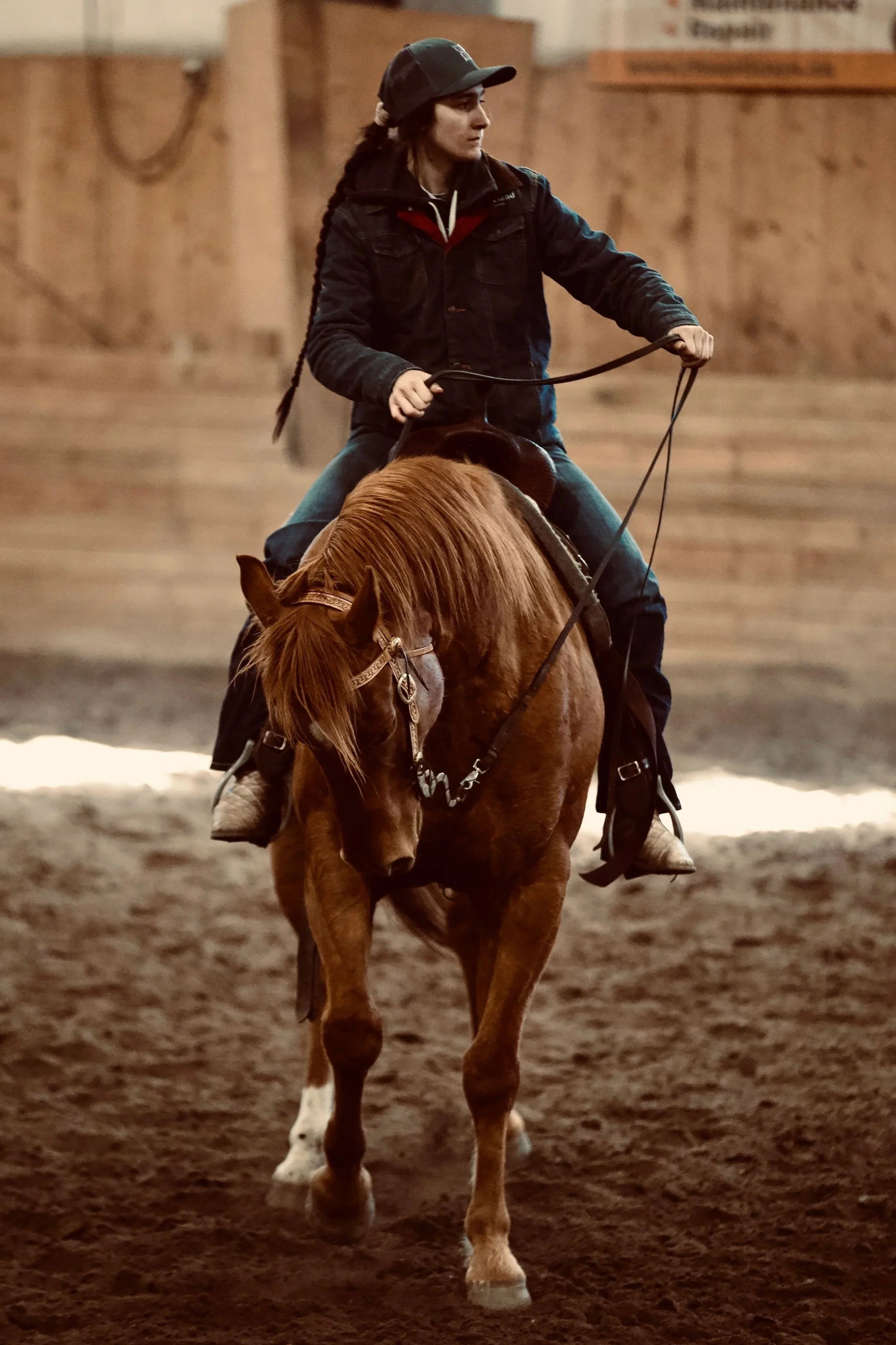A young woman riding a chestnut horse indoors, holding the reins, wearing a black cap, jacket, and jeans.