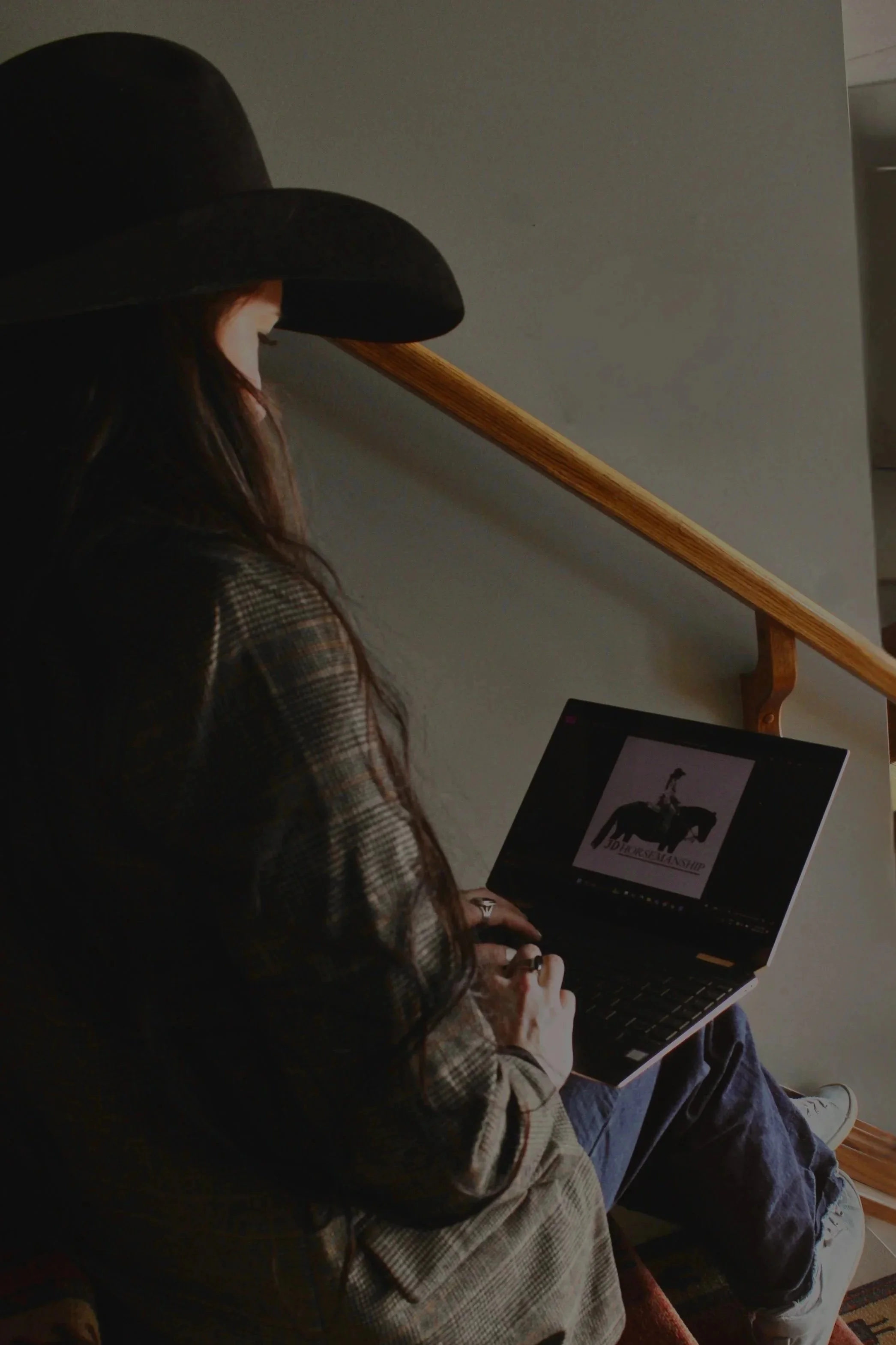A woman wearing a black cowboy hat sitting on a staircase, looking at a laptop with a graphic of a person riding a horse and the text 'JD Horsemanship' on the screen.