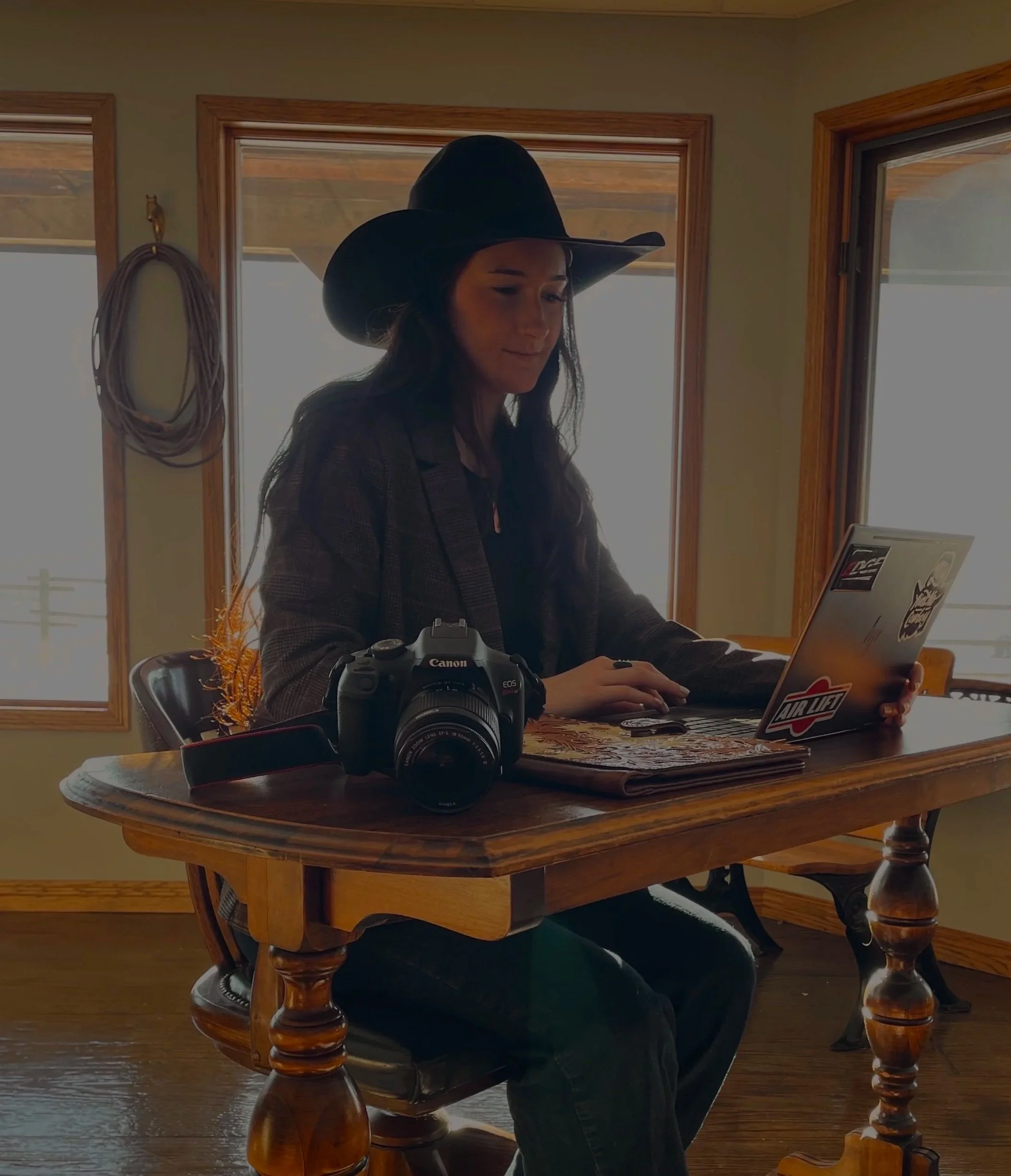 A woman wearing a cowboy hat sitting at a wooden table with a camera, a laptop with stickers, and some papers inside a room with wooden framing and windows showing an outdoor landscape.