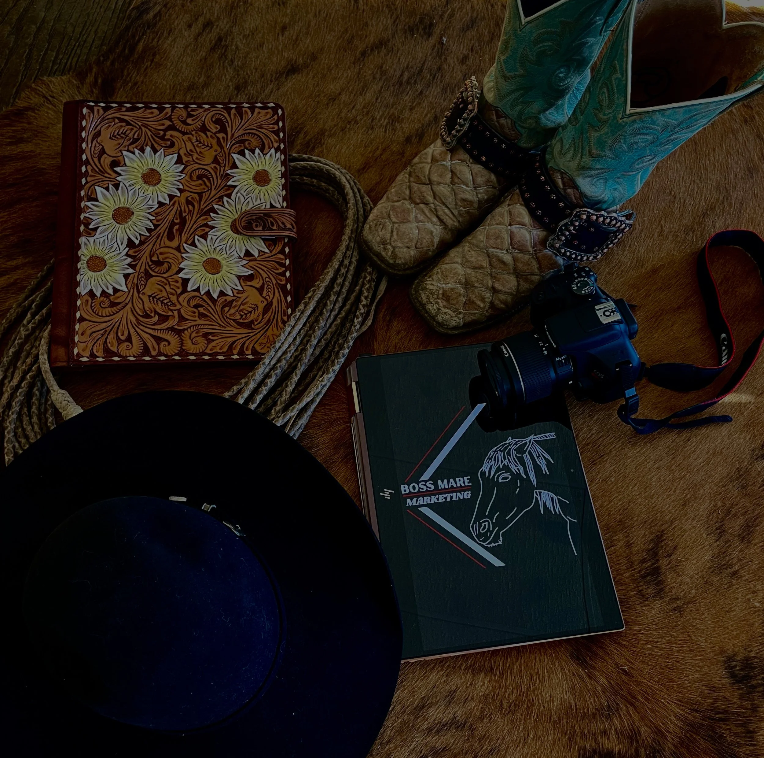 Cowhide rug with items including a black cowboy hat, a green folder with a horse illustration and the text ‘Boss Mare Marketing,’ a camera, a notebook, a pair of cowboy boots with turquoise and spurs, and a coiled rope placed on it.