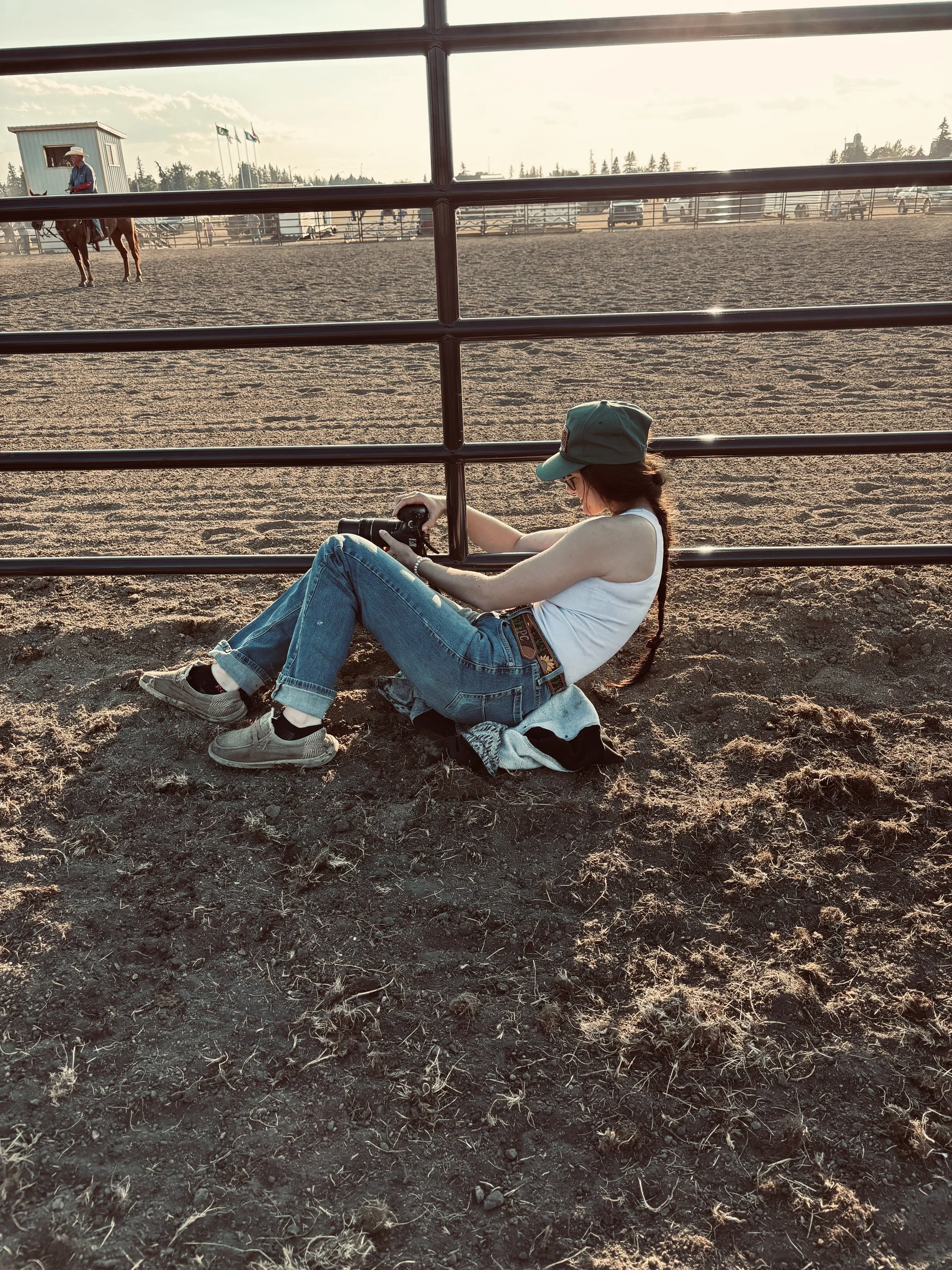 A woman sitting on the ground against a fence at a rodeo arena, holding a camera. She is wearing a cap, white tank top, jeans, and sneakers, with her hair in a braid. In the background, a person on horseback is visible, along with flags and trees under a cloudy sky.