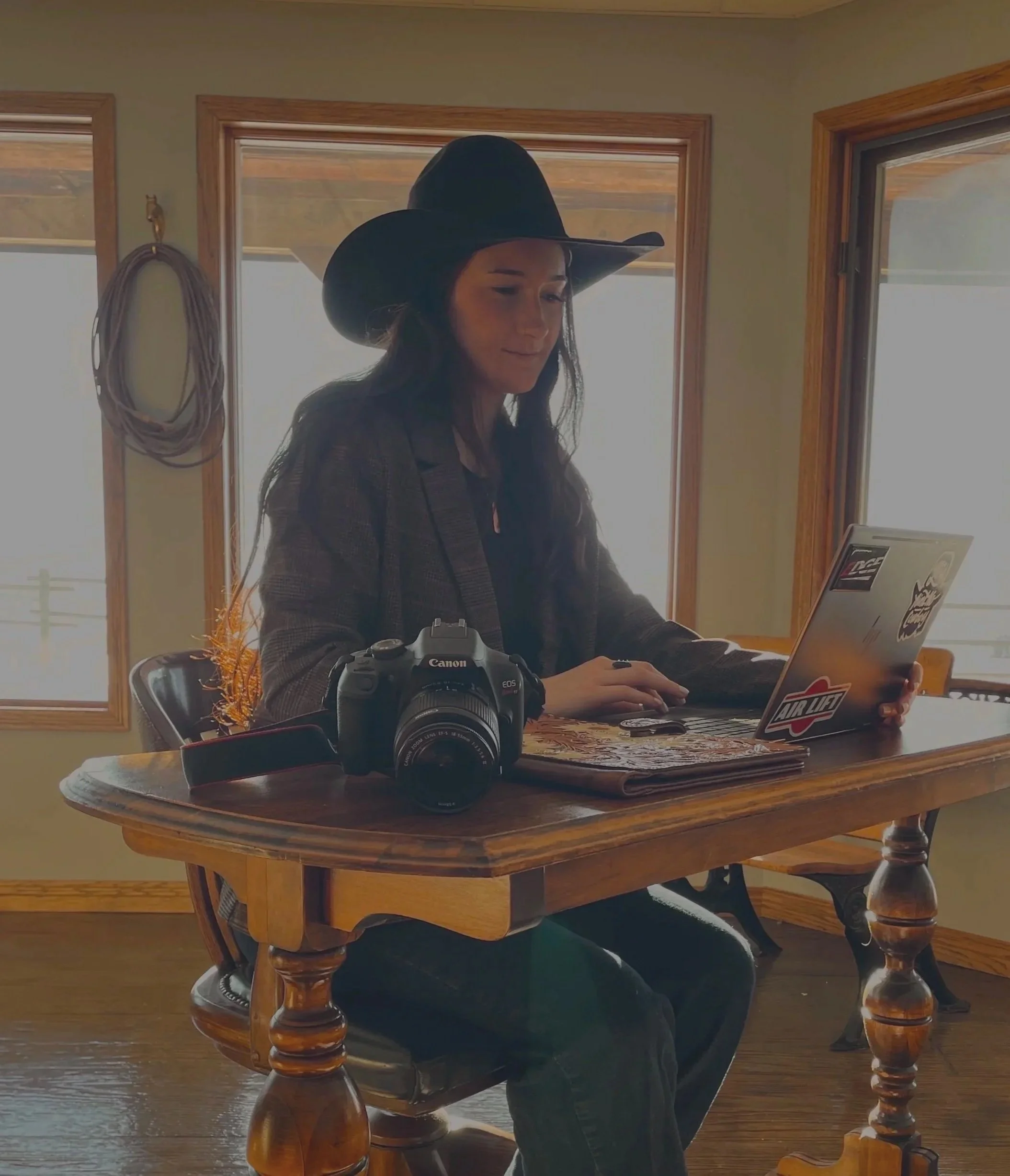 A woman wearing a black cowboy hat, plaid blazer, and jeans sitting at a wooden table with a Canon camera, a laptop decorated with stickers, and a notebook inside a room with large windows.