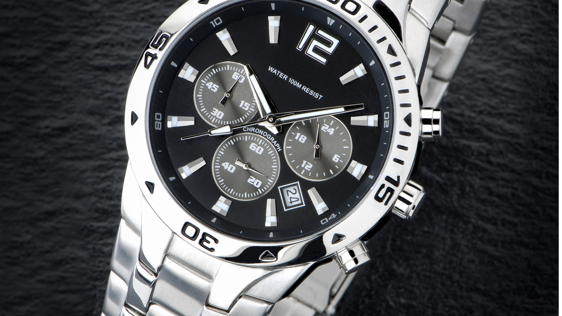 A close-up of a silver wristwatch with a black dial, three subdials, a date window at 4 o'clock, and a rotating bezel, resting on a dark textured surface.