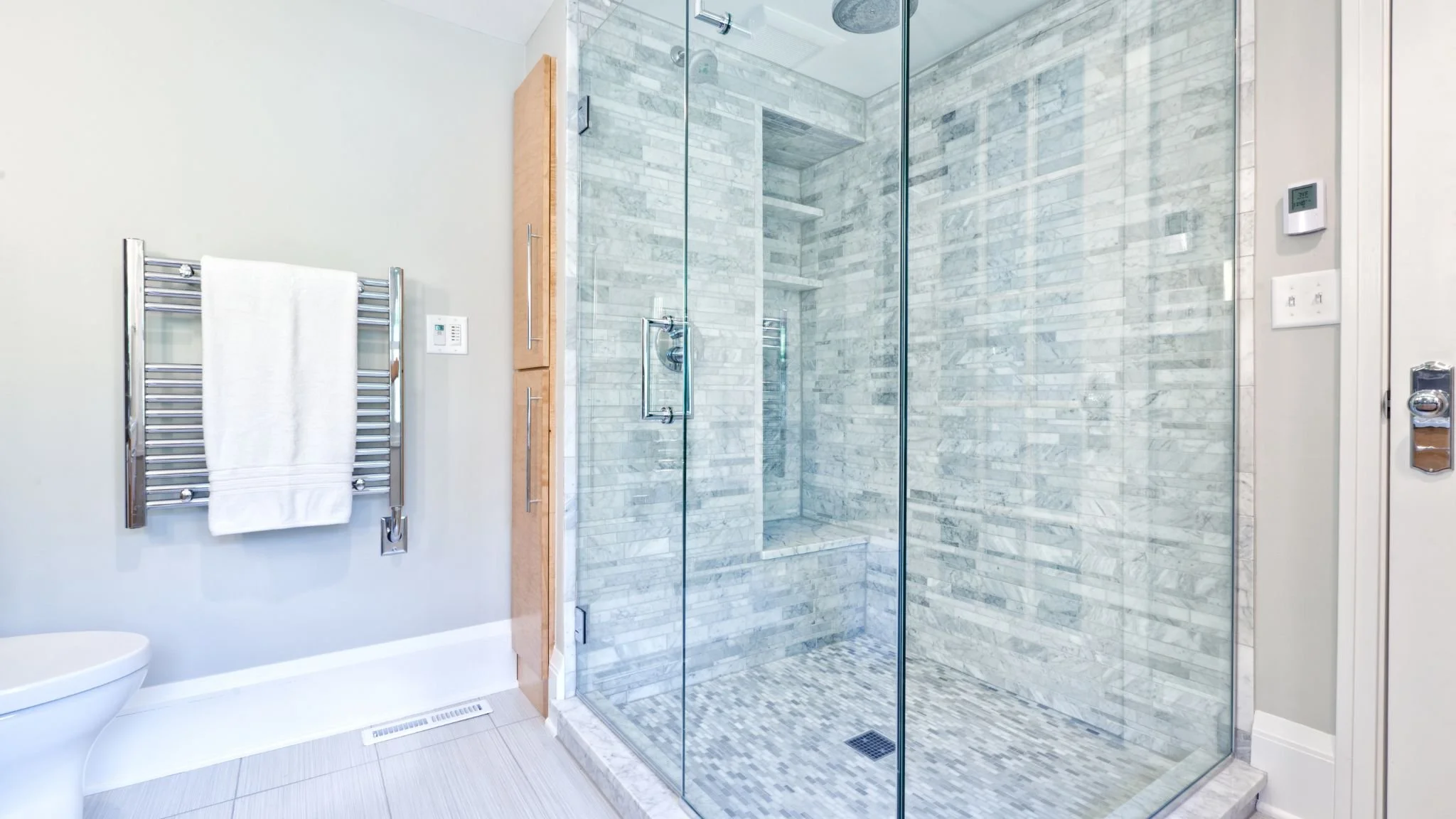 Modern bathroom with a glass-enclosed shower featuring grey marble tiles, light wood cabinetry, a towel rack with a white towel, and a white toilet in the corner.