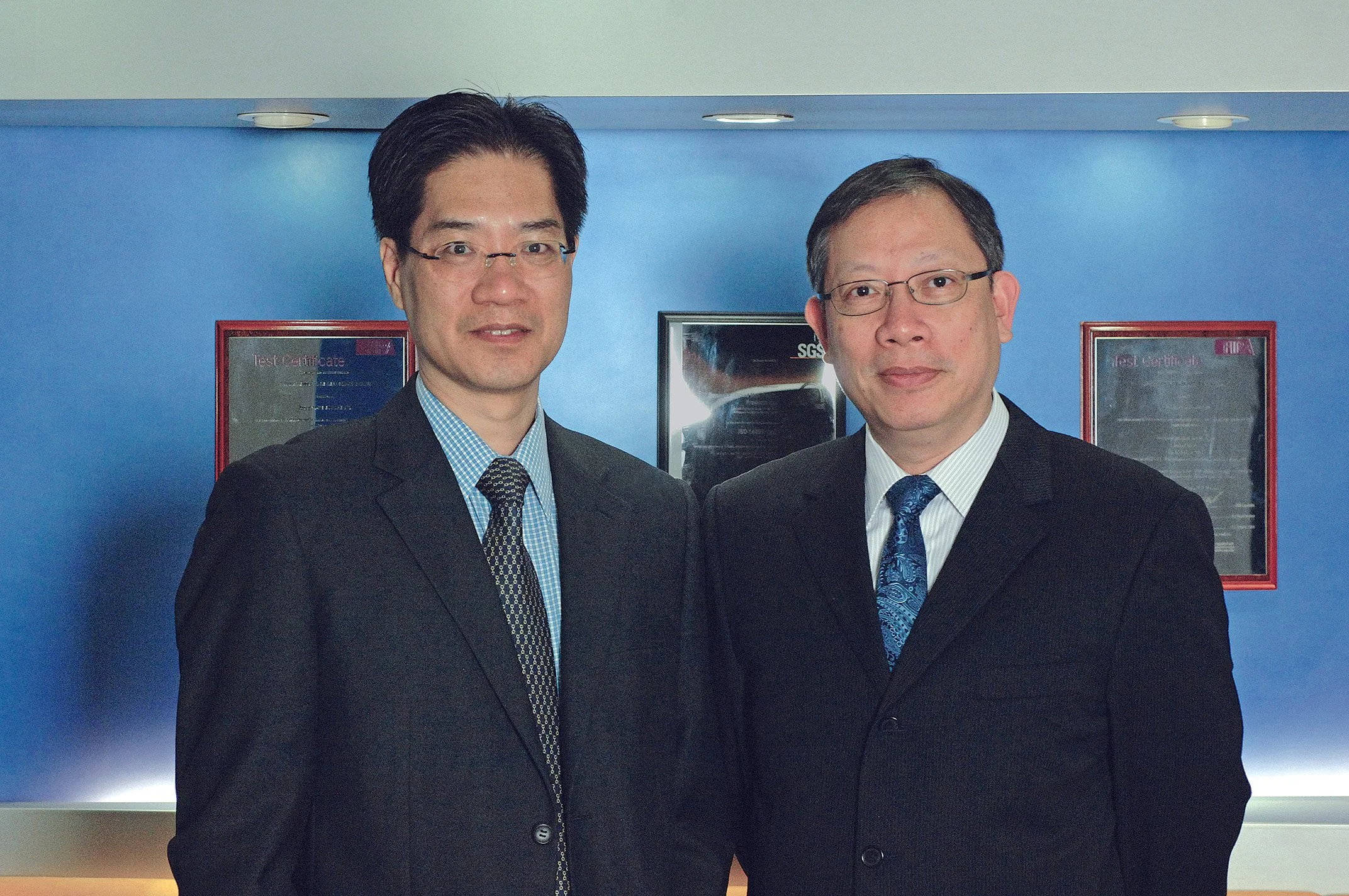 Business founders, AQUAmate business founders, Two men in formal suits standing side by side in an indoor setting with framed certificates or awards on a blue wall behind them.