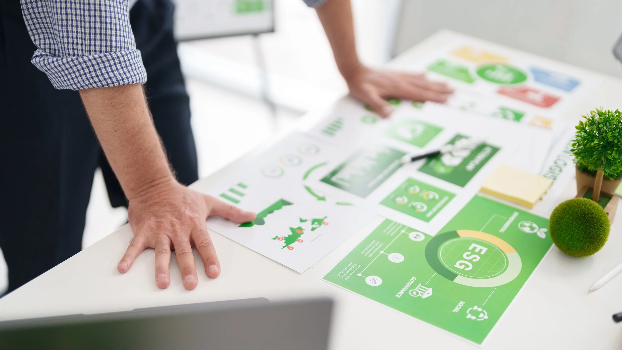 A person is leaning over a table covered with various green-colored infographic papers, charts, and diagrams related to environmental sustainability, alongside small potted plants.
