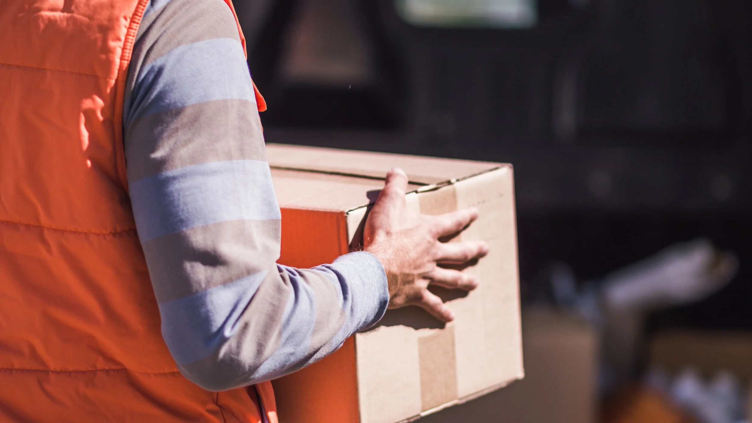 Person wearing a gray and beige striped long-sleeve shirt and an orange vest holding a cardboard box outdoors near a black vehicle.