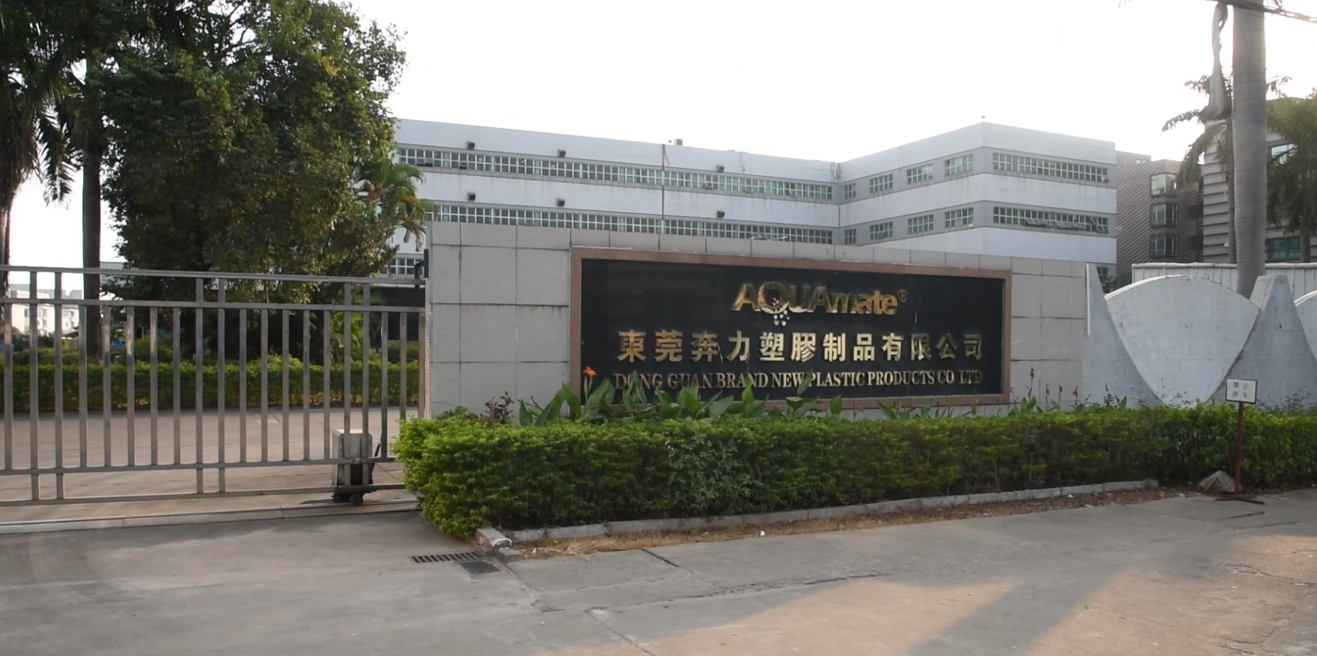 Entrance gate with a sign for Dong Guan Brand New Plastic Products Co Ltd, featuring trees, bushes, and a large white building in the background.