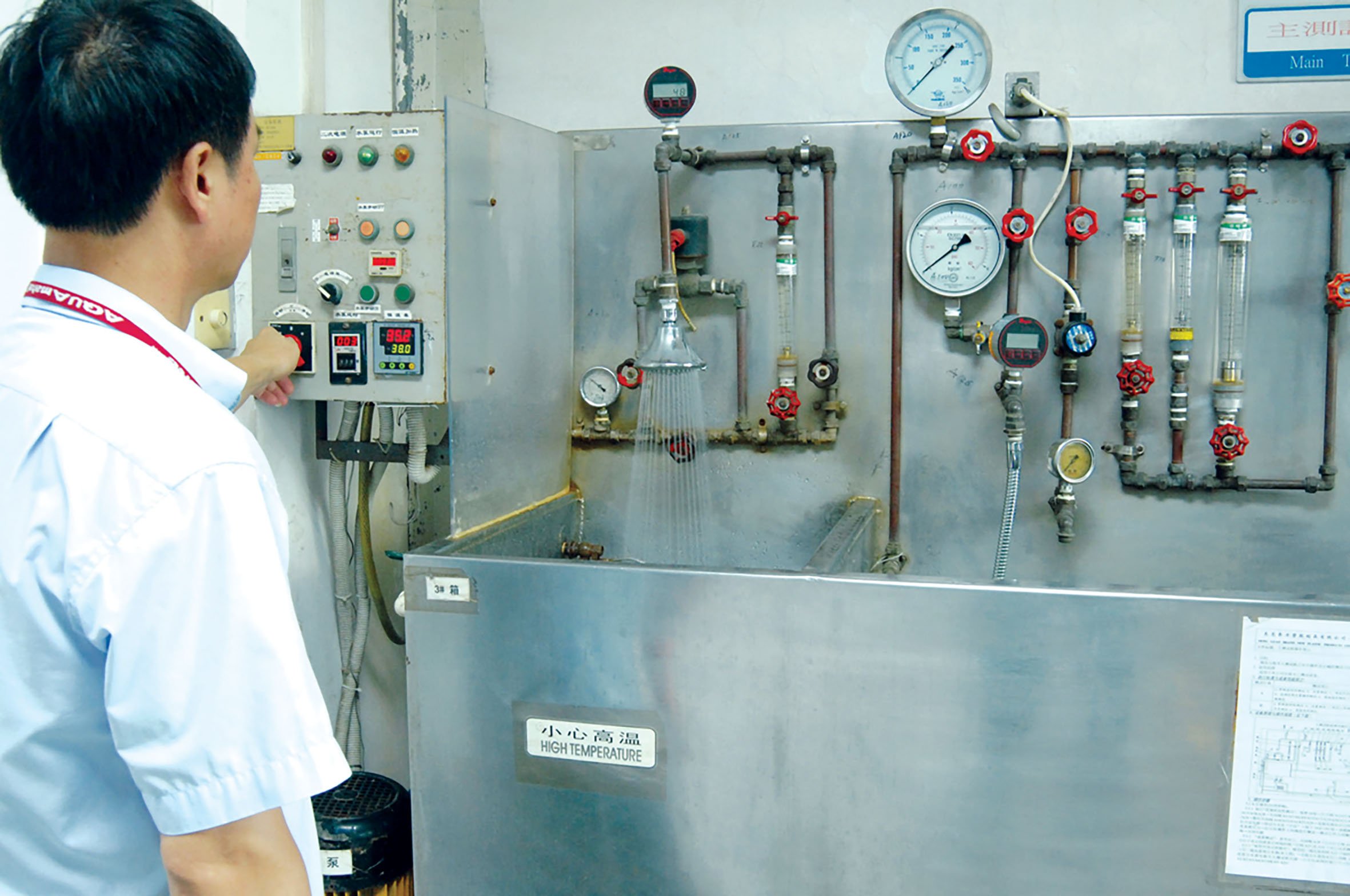 A man adjusting controls on an industrial water boiler system with gauges, valves, and pipes. The boiler has a sign that reads 'High Temperature' in both Chinese and English. Testing the quality of showerheads