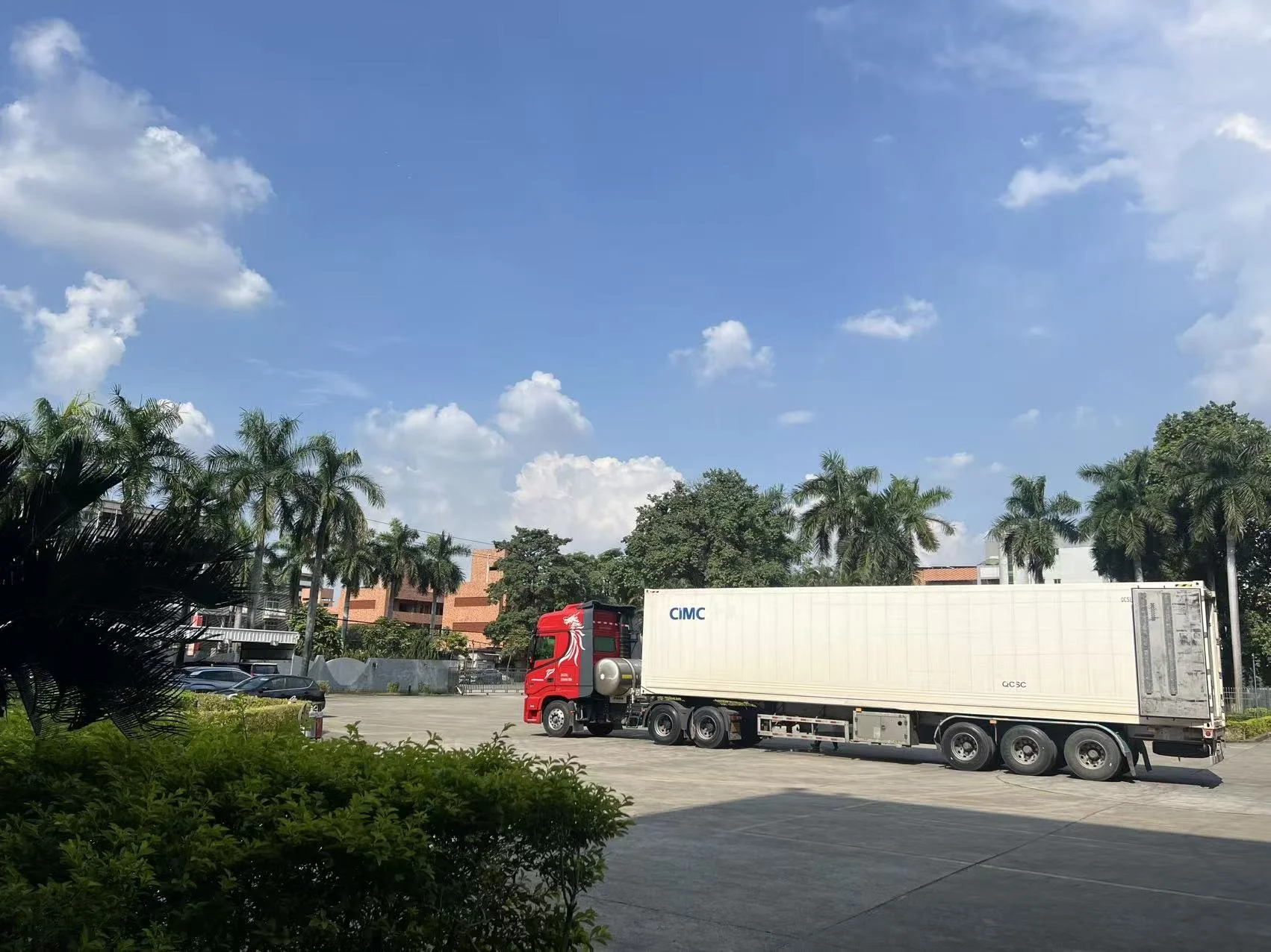 Red semi-truck with a white trailer parked on a concrete lot, with palm trees and a blue sky with clouds in the background.