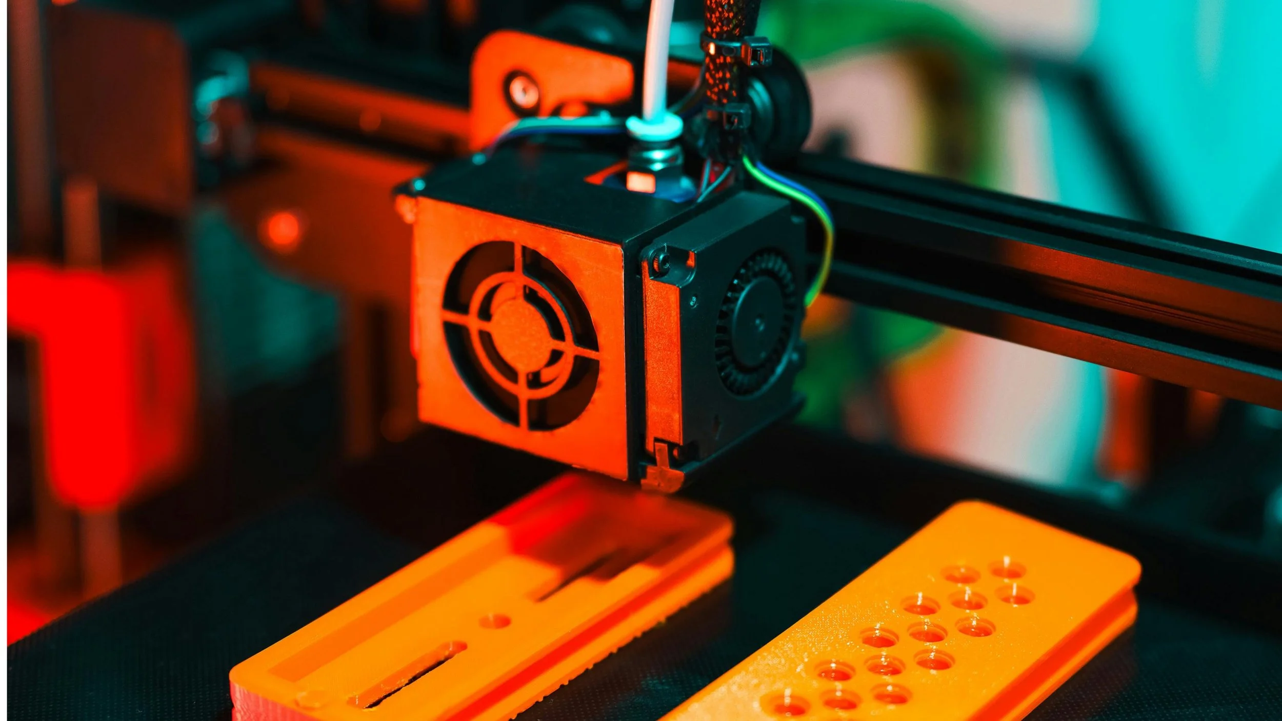 Close-up of a 3D printer extruder printing with orange filament, with orange printed parts underneath on a textured black surface, illuminated with colorful lighting.