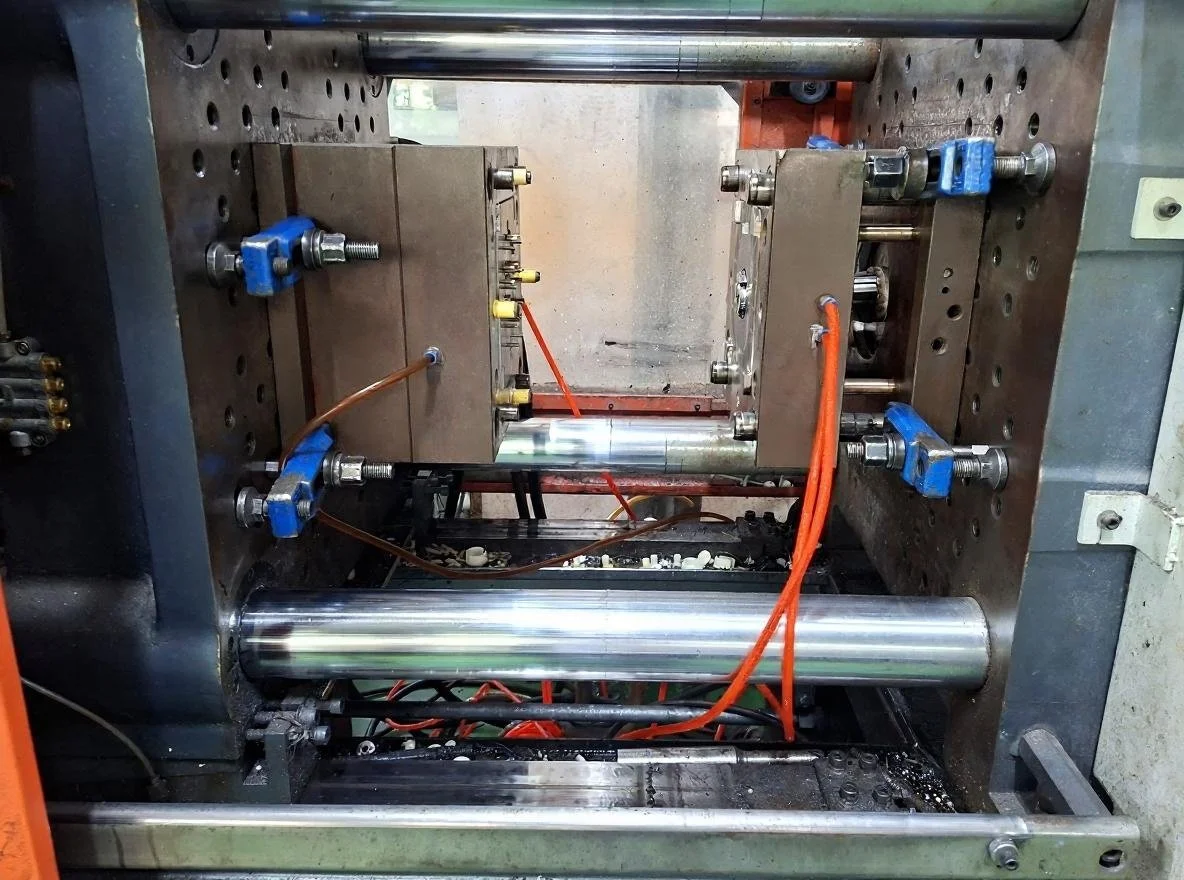 Close-up view of a mechanical rig with metal cylinders, orange and blue wiring, and metal plates with bolt attachments, inside a workshop or industrial setting.