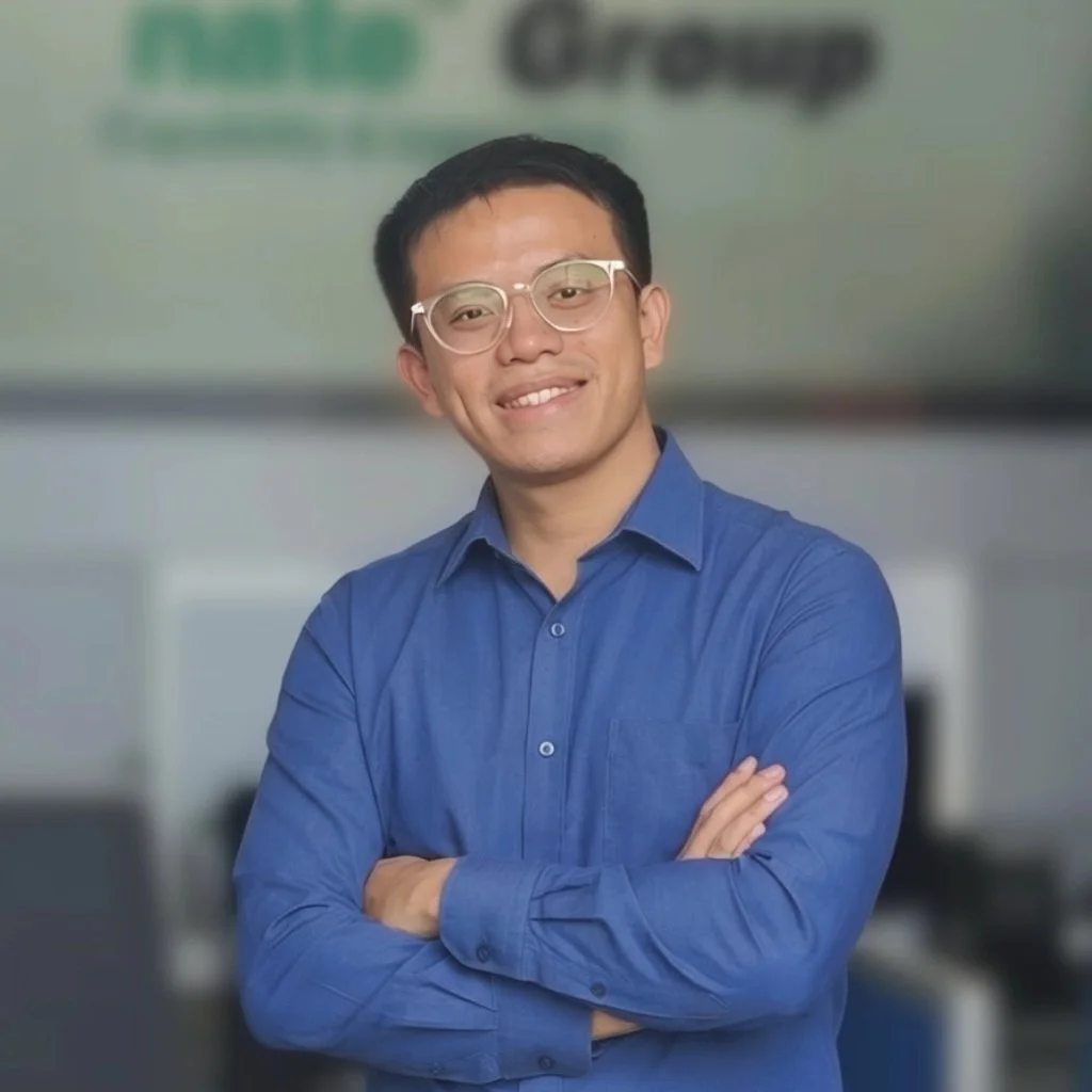 A man with short black hair and glasses smiling at the camera, wearing a blue button-up shirt, standing with arms crossed in an office setting.