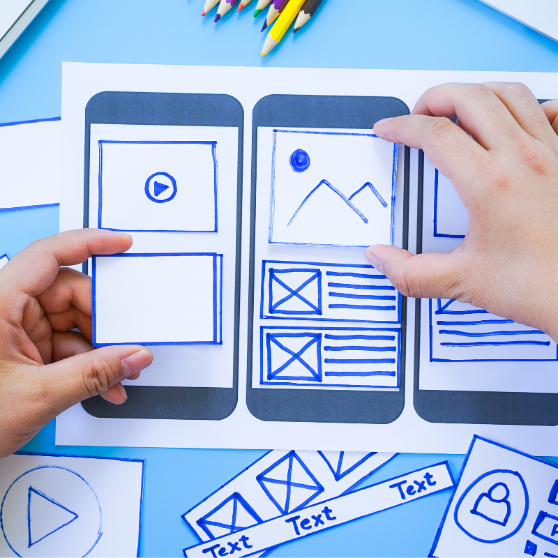Hands sketching wireframes of a mobile app interface with sketches of video, image, and text sections, surrounded by colored pencils and additional paper sketches with icons and text.