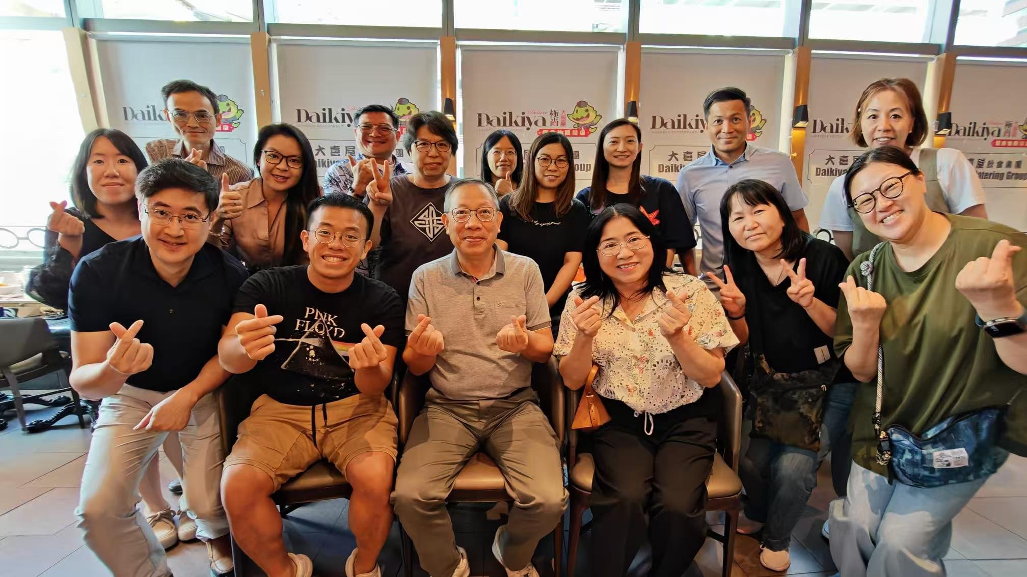 Group of people gathering indoors for a photo, with a backdrop featuring the 'Daikiya' logo and a cartoon frog mascot, smiling and making finger heart gestures.