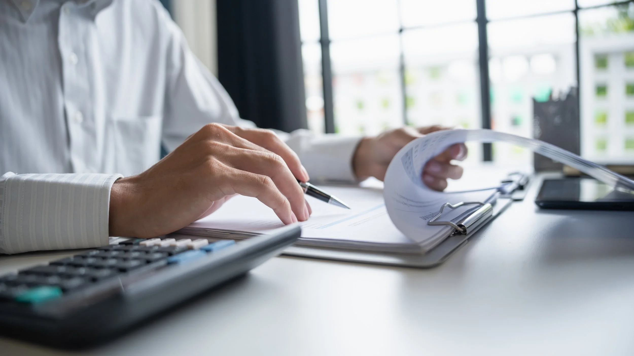 Person in white shirt reviewing documents at desk with calculator, pen, and clipboard, near large window