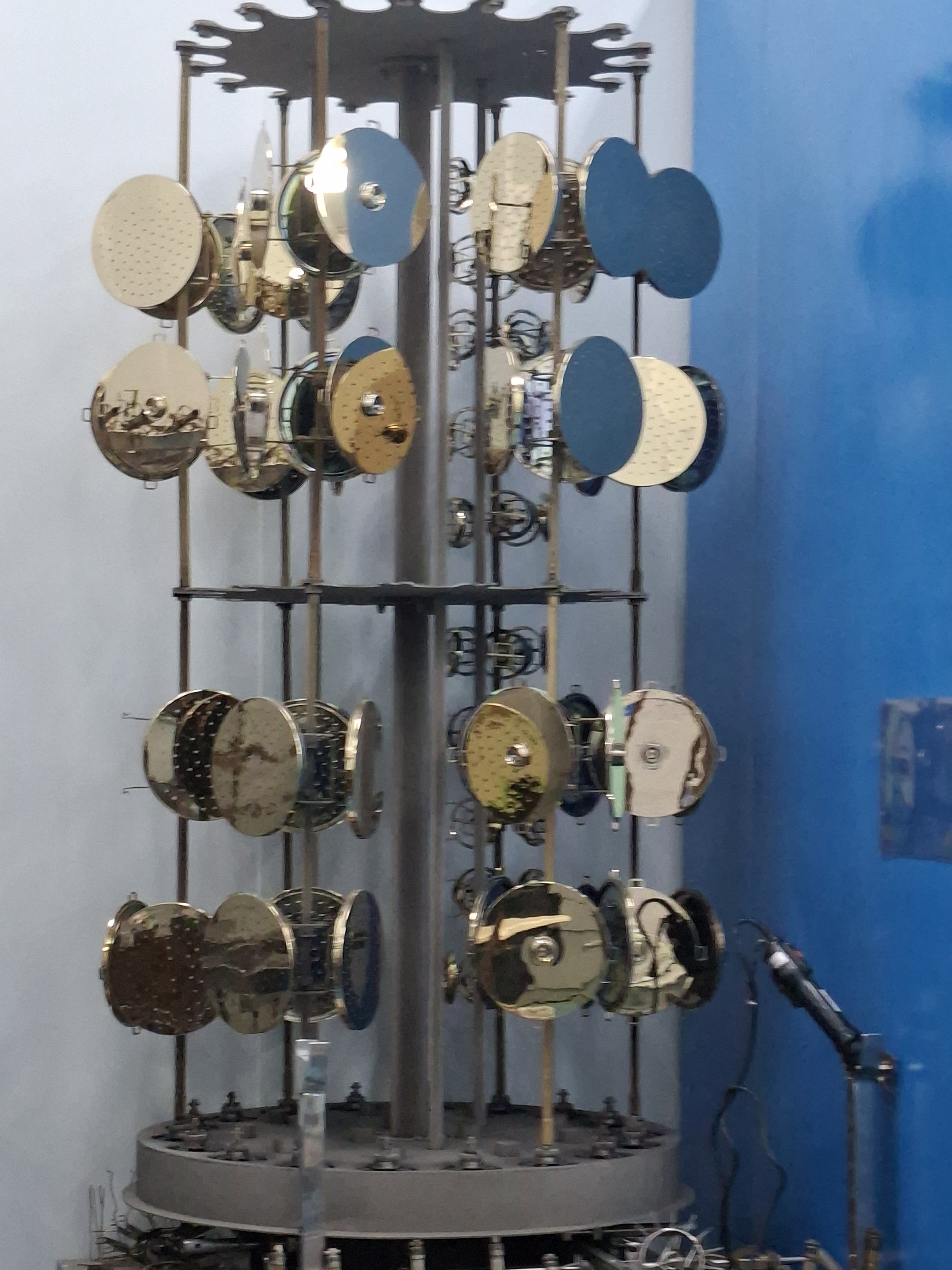 A large, vintage, mechanical clock mechanism with multiple golden and dark-colored gears and wheels, mounted on a vertical metal frame against a blue wall.