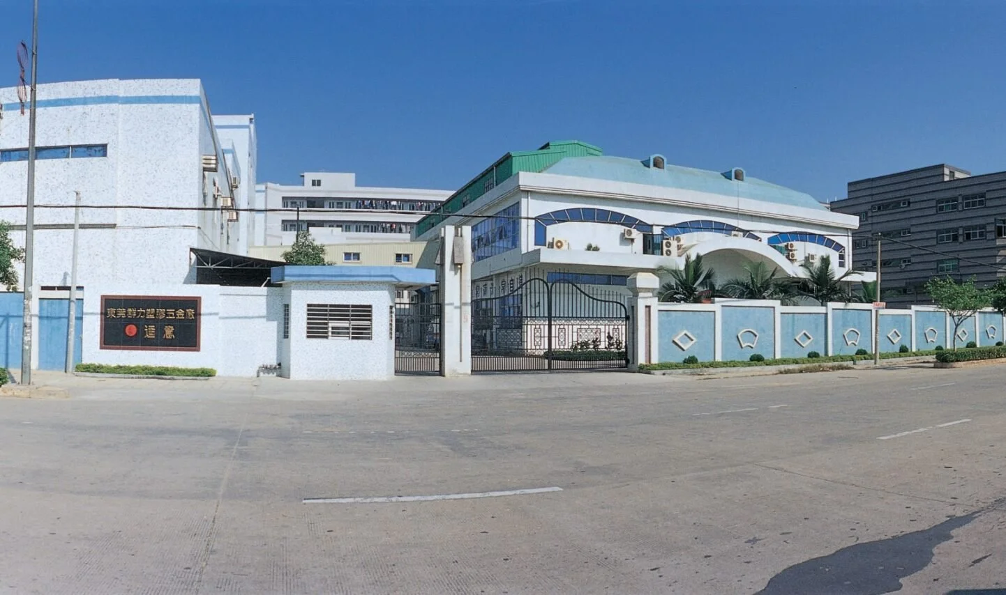 A large, white building with blue accents, a green roof, and decorative fences, situated in an urban area with other tall buildings in the background. There is a black gate at the entrance and a sign with Chinese characters on the wall.