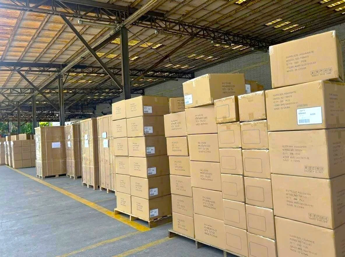 Stacks of cardboard boxes arranged on pallets in a warehouse with a metal roof and open sides.