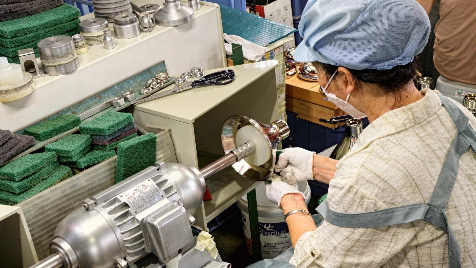 A woman wearing a face mask, gloves, and a blue cap is polishing a metal object with a polishing machine in a workshop. There are various tools, sponges, and metal parts on the work surface.